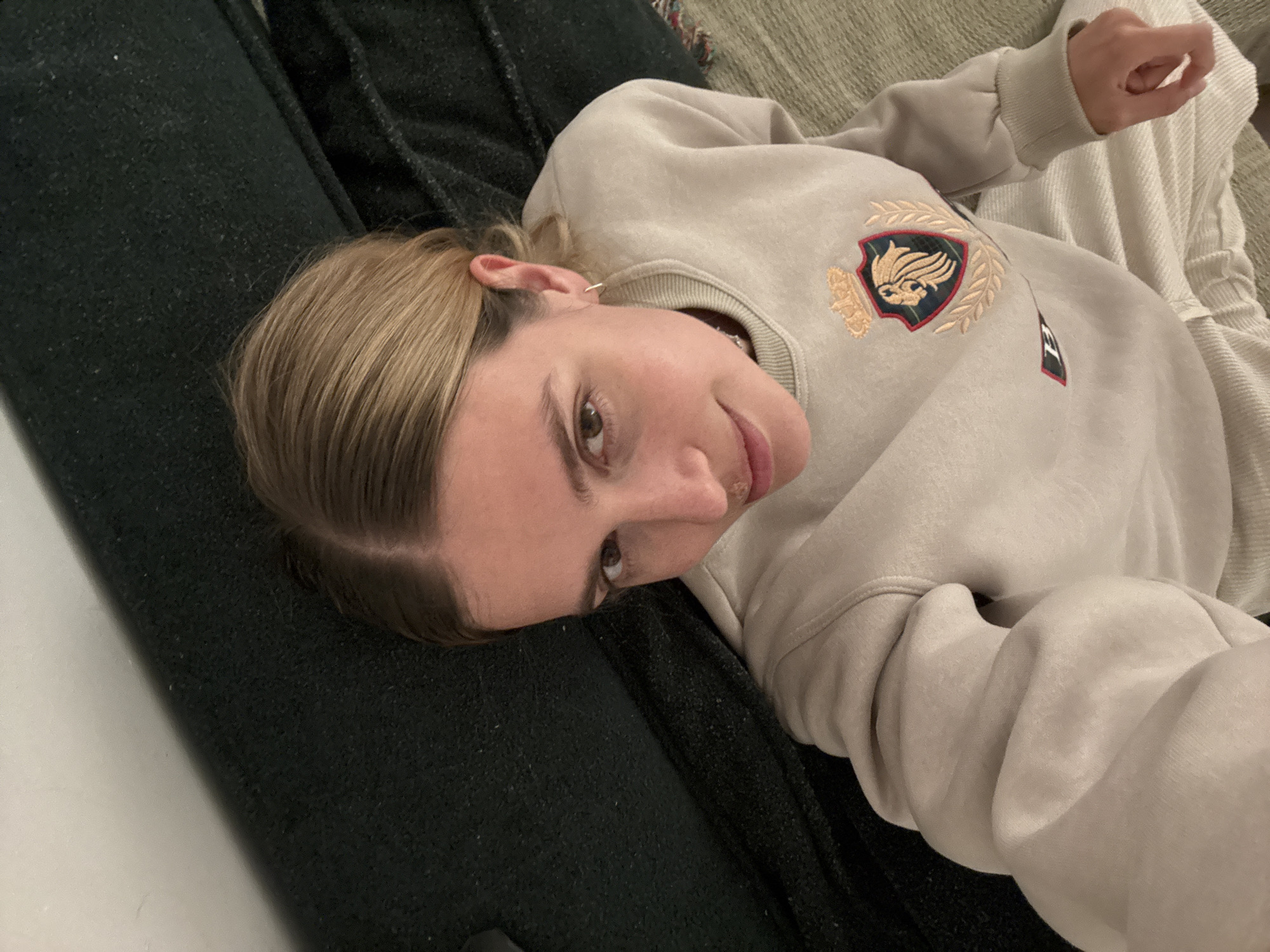 A person is reclining on a dark-colored couch. They are wearing a light-colored sweatshirt with an embroidered crest. The location is Unknown, Unknown.