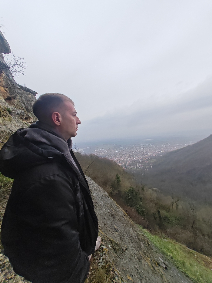 A male individual is depicted in profile, facing right, standing on a rocky ledge overlooking the city of Shumen, Bulgaria. The vantage point is an elevated natural feature, consistent with the Shumen Plateau, with a rugged, light-colored rock face to the subject's immediate left. The individual is wearing a dark winter jacket over a grey hooded sweatshirt. His gaze is directed towards the panoramic cityscape below, which shows densely packed buildings extending through a valley. The city is flanked by hills covered in deciduous trees, largely devoid of foliage, suggesting a late autumn or winter season. The sky above is uniformly overcast and grey, providing diffused lighting to the entire scene. The rocky ledge itself is uneven, grey, with minor patches of green vegetation.