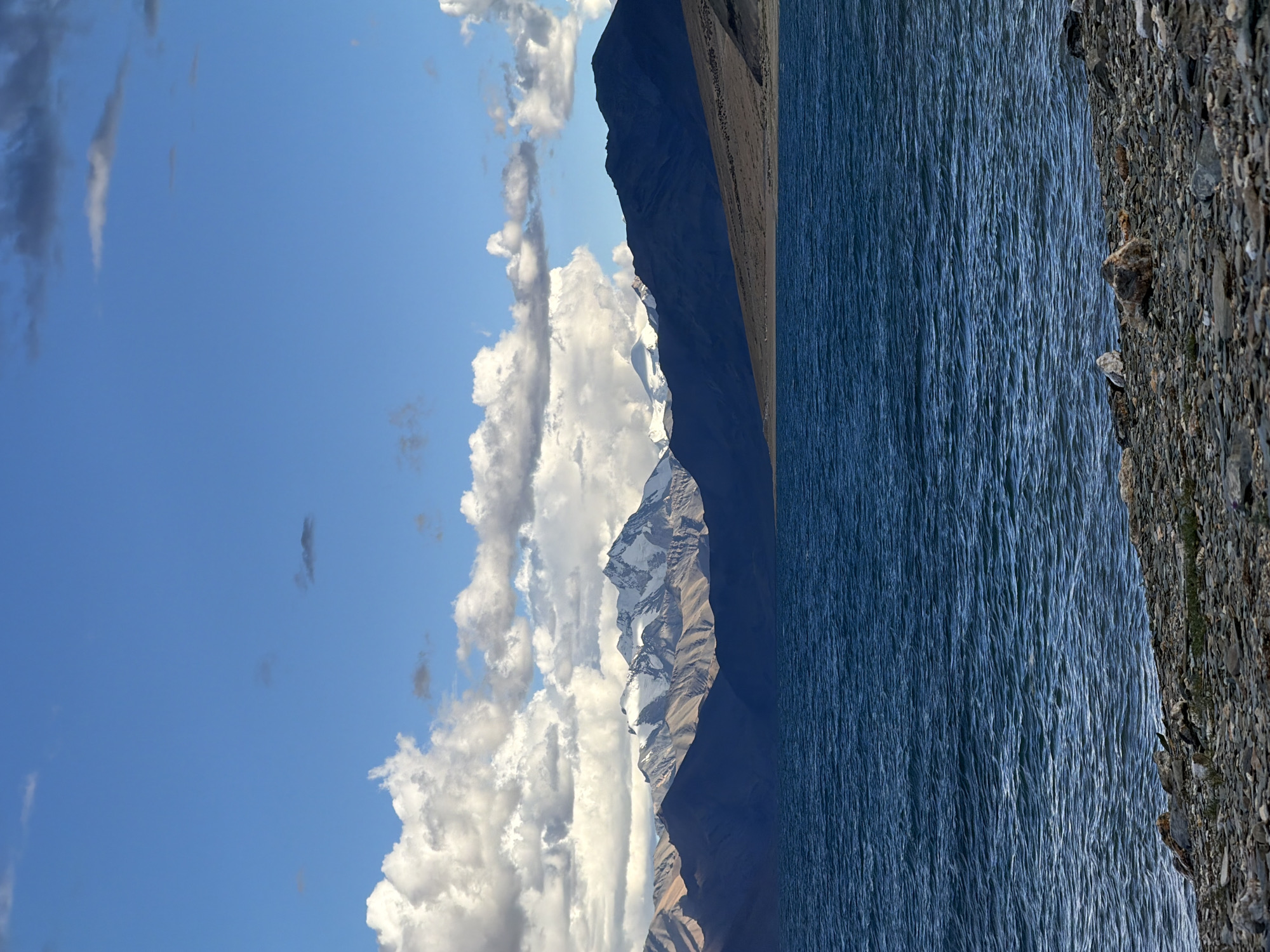 The image depicts a serene natural landscape featuring a body of water, mountains, and a sky filled with clouds. In the foreground, a rocky shore borders the dark blue water, which is rippled by a gentle breeze. Beyond the water, a range of mountains stretches across the horizon. The lower slopes of the mountains are a muted brown and beige, transitioning to darker, shadowed forms as they recede. The peaks of the mountains are snow-capped, gleaming white against the sky. The sky itself is a vibrant blue, punctuated by large, fluffy white clouds that cast shadows on the mountain ranges. The overall scene conveys a sense of natural beauty and tranquility, likely in a high-altitude region. The location is identified as being in the city of Unknown, India.