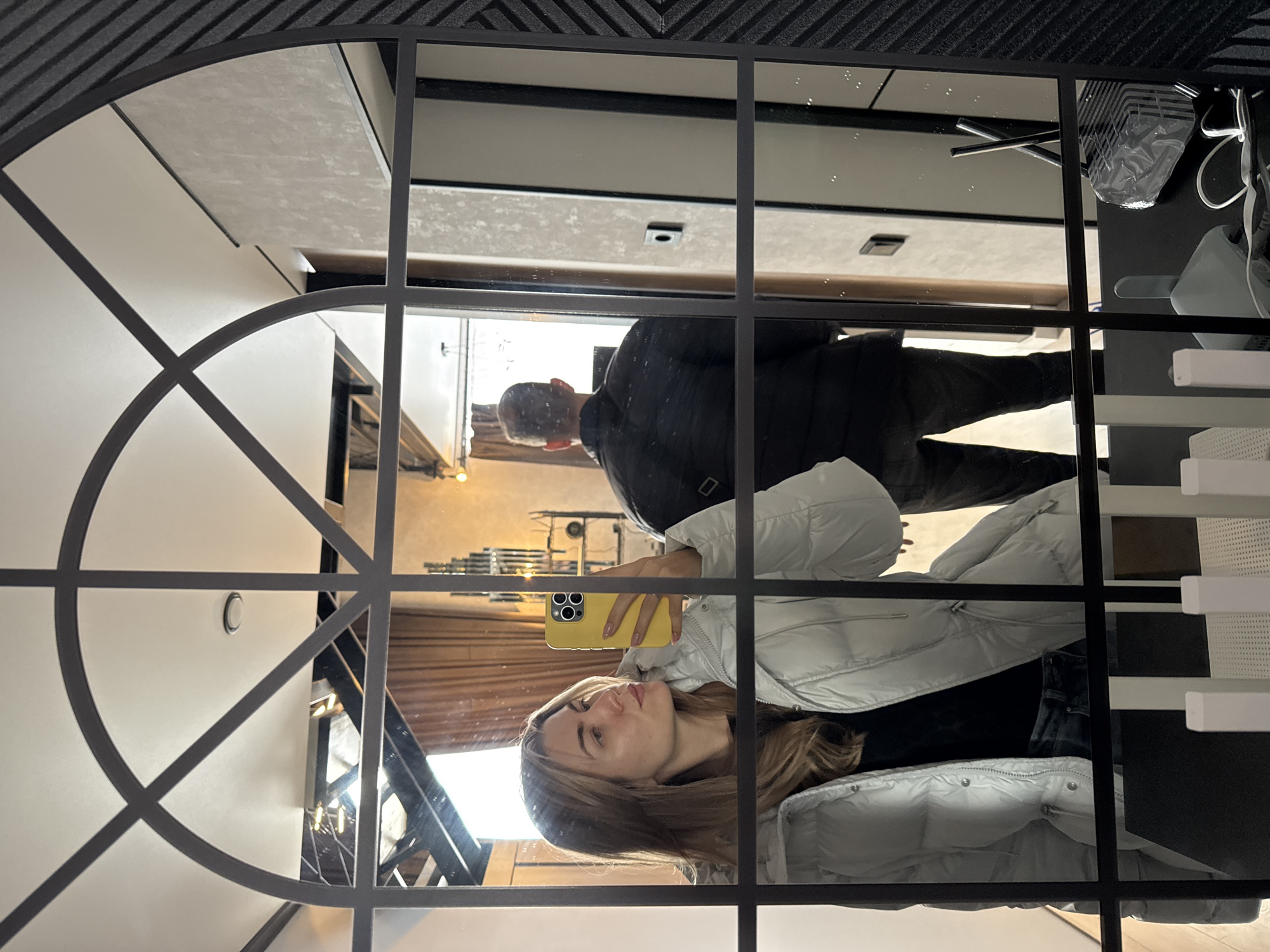 The image displays a mirror selfie taken within a modern indoor setting. A prominent, large mirror with a distinctive black frame, featuring both straight lines and elegant arched segments, is central to the composition.
Reflected in the mirror, a young woman takes the photograph, holding a bright yellow smartphone. She has long, light brown hair and wears a light grey puffer jacket over a dark top, with a neutral expression. To her upper right, the reflection of a man is partially visible, wearing a dark, possibly quilted or plaid, jacket.
The reflected environment suggests a contemporary interior. On the left, a light-colored wall features a circular light fixture. Further back, warm lighting highlights architectural elements, possibly stairs or railings, and wooden textures. On the right side, a dark, textured ceiling or wall is visible, possibly adorned with acoustic panels. Below this, a white and grey shelving unit contains items such as a light grey shoe and other belongings. Metallic elements, possibly part of a chair, are also reflected. The scene depicts a well-lit, stylish indoor space. No visible text is present.