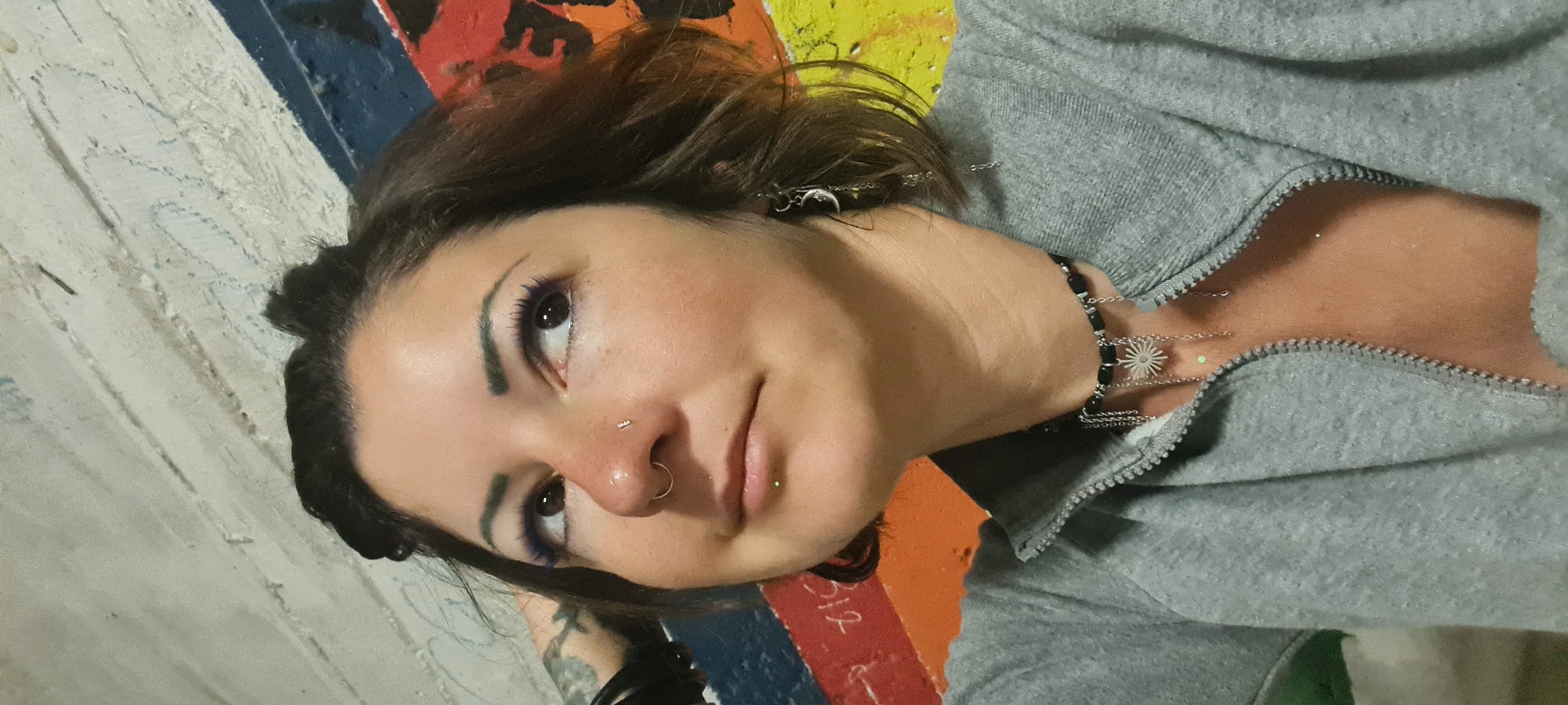 A woman is positioned on a painted surface featuring multiple colors. She has brown hair and brown eyes with purple eyeliner. She has piercings in her nose and lip, along with earrings. She is wearing a gray hooded sweatshirt and a necklace with multiple chains and a starburst charm. Her left arm appears to have a tattoo. The location is not specified as Rishon LeZion, Israel, in the image, therefore, the description cannot confirm this as the location.