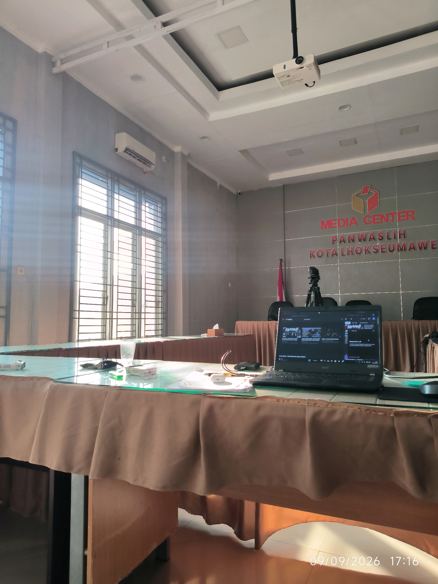 The image depicts the interior of a room, likely a meeting or media room, within the "MEDIA CENTER PANWASLIH KOTA LHOKSEUMAWE" in Lhokseumawe, Indonesia. The room is equipped with a projector mounted on the ceiling, air conditioning units on the wall, and large windows with blinds. A table covered with a brown cloth is prominent in the foreground, with a laptop and other items placed on its glass surface. The laptop screen displays content that suggests it is in use. The overall environment appears to be a professional setting.