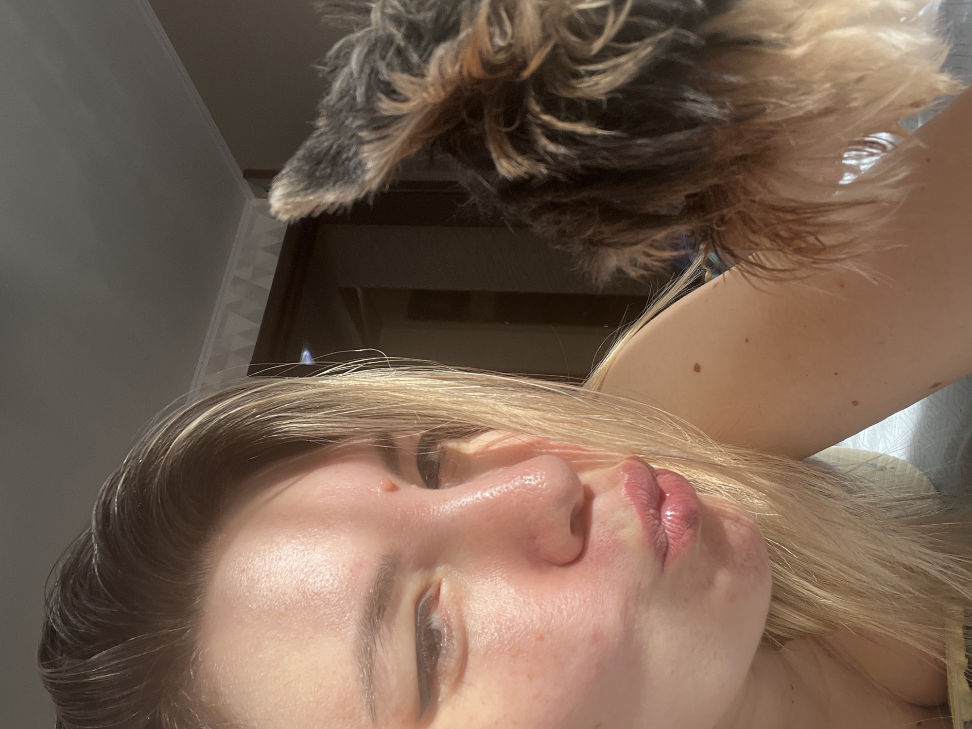 The image is a close-up selfie featuring a young woman with blonde hair, pouting her lips and looking towards the right. She has fair skin with some blemishes and wears subtle eye makeup. To the right of the woman, a fluffy dog's ear and back are visible, with fur exhibiting shades of brown and gray. The background is dimly lit, showing a portion of a wall with decorative molding and a darker area that appears to be a doorway or furniture. Sunlight casts a warm glow on the woman's face, particularly on her forehead and cheek. The scene suggests an indoor setting, likely a bedroom or living room. There is no discernible information in the image that would indicate its location in the city of Sochi, Russia.