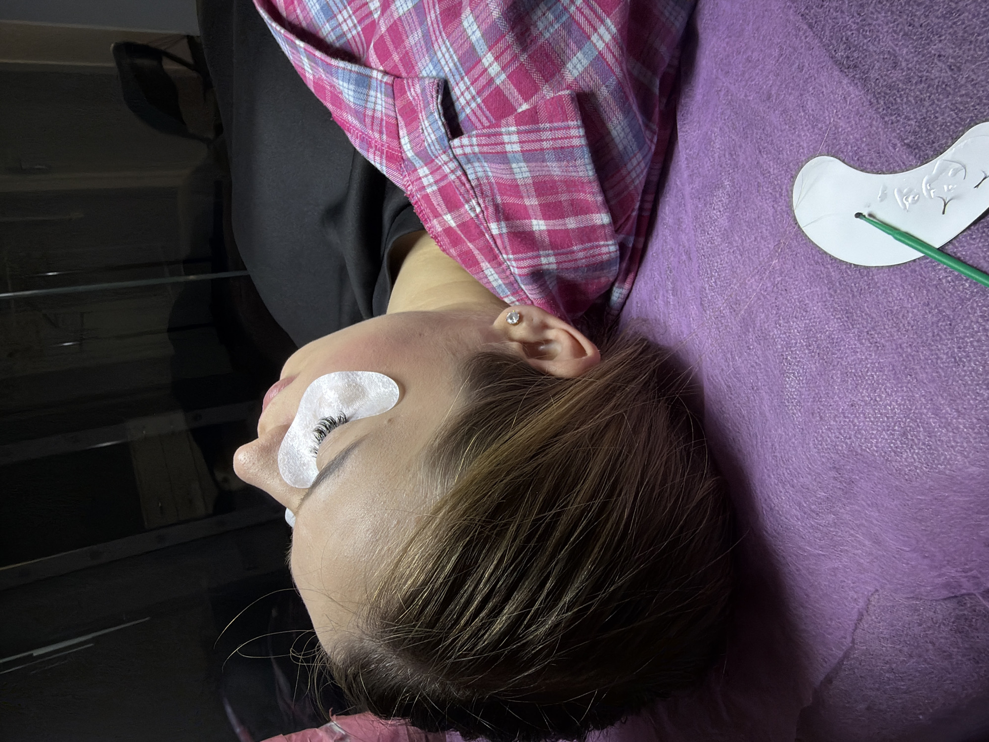 A young woman with light brown hair is lying on her back on a purple textured surface, likely a bed or treatment table. She is wearing a pink and blue plaid shirt and has white eye patches under her eyes, indicating she is undergoing a beauty treatment, possibly eyelash extensions. Her eyes are closed, and she appears relaxed. The background is dark and out of focus, showing hints of furniture and reflections. To the lower right, a white, curved tool with a green applicator is resting on the purple surface. The scene suggests a professional beauty salon or a home setting where such treatments are performed. The location is identified as Kyiv, Ukraine.
