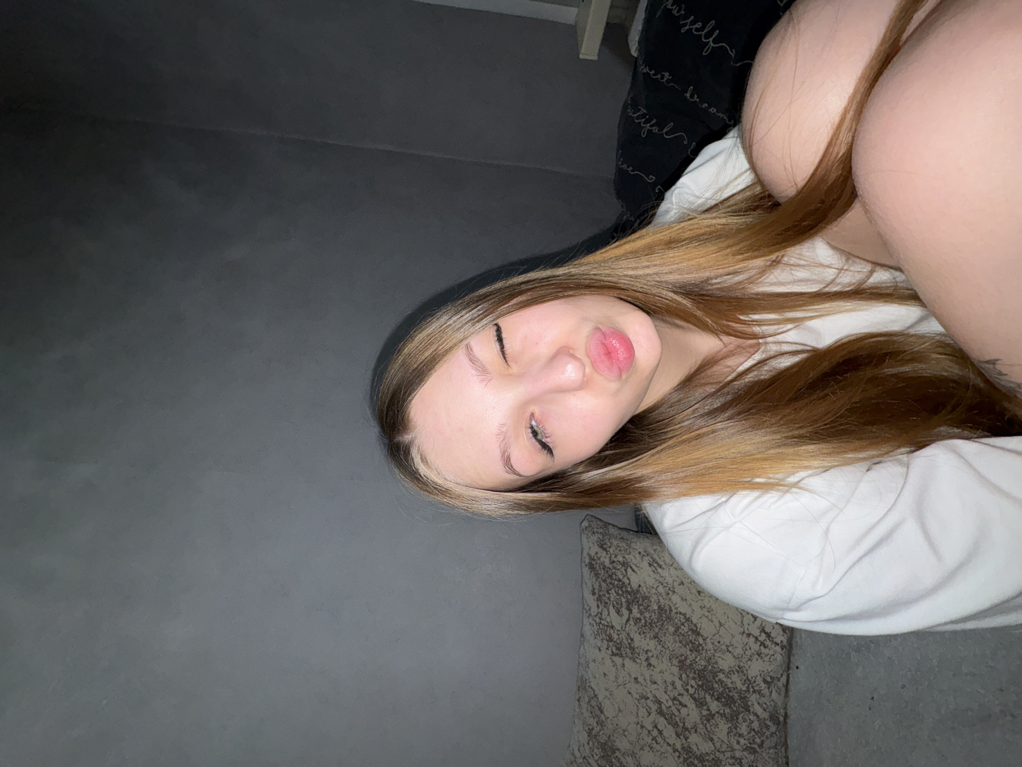 A young woman with long, light brown hair is lying down. She is wearing a white shirt. Her eyes are closed, and she is making a kissing face. The background is a gray wall, a patterned pillow, and a dark object with white writing. The location is not discernible from the image.