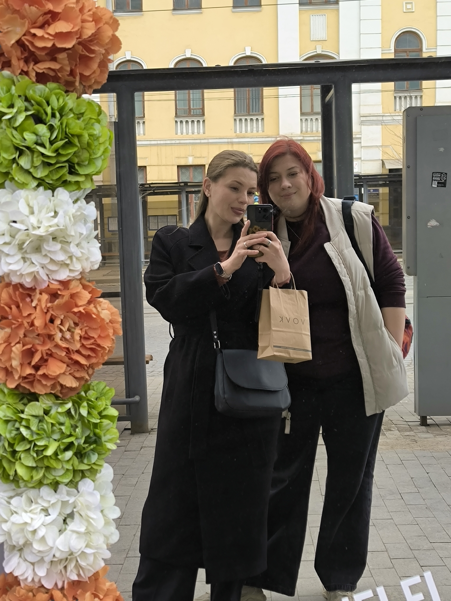 Two women are reflected in what appears to be a bus stop shelter with decorative floral arrangements. The woman on the left has light blonde hair, is wearing a black coat, and holds a smartphone, taking a selfie. She also wears a black shoulder bag. The woman on the right has reddish-brown hair and wears a dark burgundy sweater, black trousers, and a light beige vest. She is carrying a brown shopping bag with the text "AVOV" printed on it. They stand on a paved surface with gray patterned tiles. Behind the bus stop, a yellow and white building with rows of windows is visible. This is in Vinnytsia, Ukraine, based on the AVOV bag. The lighting suggests it is daytime, under an overcast sky.