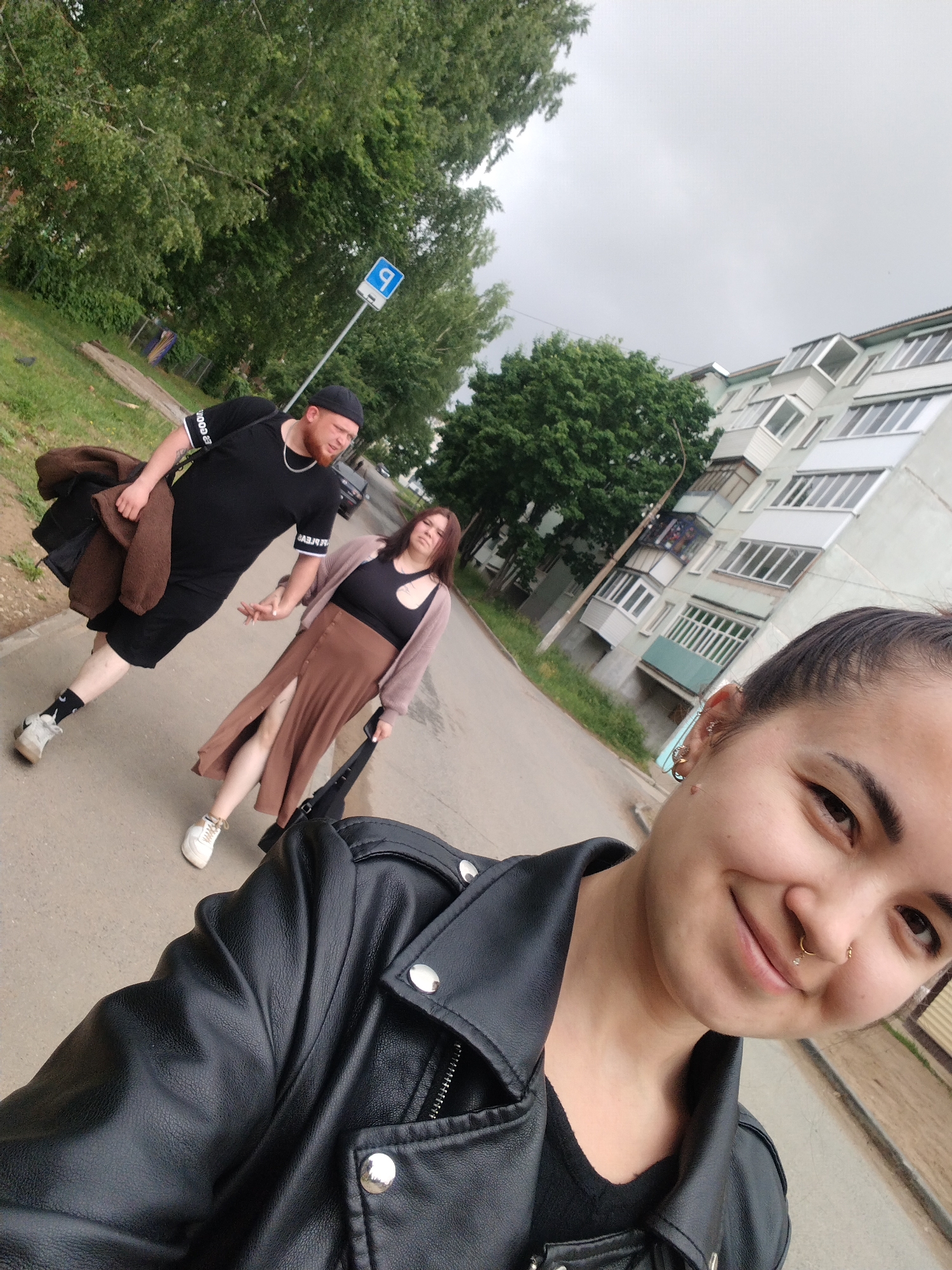 This outdoor selfie captures three individuals on a street in Noviy, Russia, under an overcast sky. In the immediate foreground, a young woman with dark hair tied back smiles directly at the camera. She is wearing a black leather jacket over a black top and has a nose ring and several ear piercings.
Behind her, walking down a paved path, are two other people. On the left is a man with a reddish beard, a black beanie, and a black t-shirt. The t-shirt's sleeves have white trim, with "GOOD" visible on one sleeve and mirror-imaged text on the other. He wears black shorts, patterned white socks, and white sneakers, and carries a brown jacket or blanket over his arm. Beside him, a woman with long brown hair wears a black top, a long brown or rust-colored skirt with a front slit, a light-colored cardigan, and white sneakers. She carries a black bag in her right hand. The man appears to be looking towards the camera.
The background features green trees and foliage alongside the path. A multi-story apartment building with numerous balconies and windows is prominent on the right. A blue square street sign with a white symbol, possibly a stylized 'P' for parking or an arrow, is visible on a pole amidst the trees. The sky is uniformly grey and cloudy, suggesting a cool or mild day. The image has a casual, everyday feel.