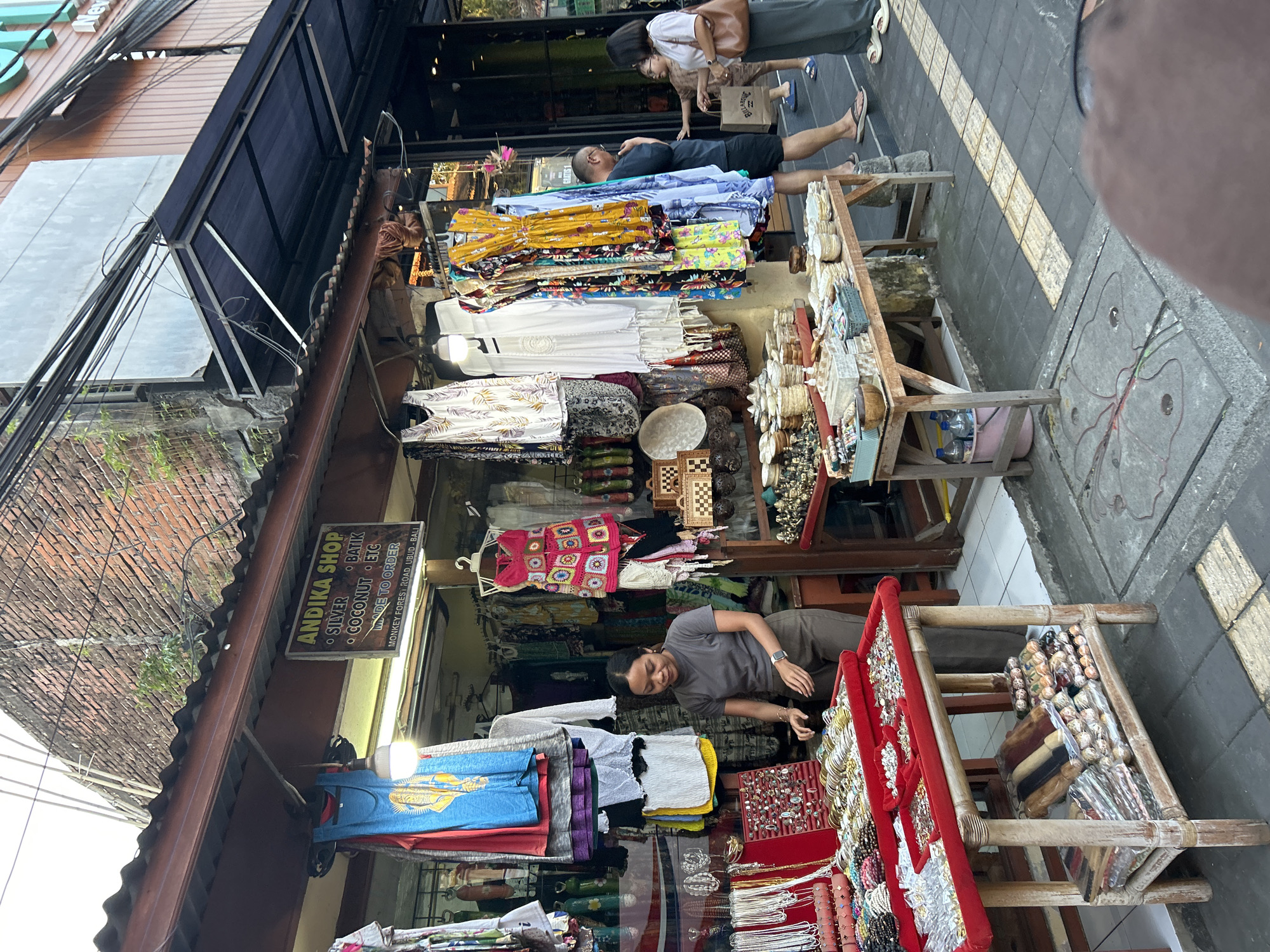 The image shows a street scene in Ubud, Indonesia, with a shop called "ANDIKA SHOP" displaying various goods, including clothing and accessories. A woman in a grey t-shirt and brown pants is standing behind a table filled with jewelry, appearing to be a vendor. Several customers are browsing the items, including a man in a black t-shirt and shorts and a woman in a white shirt and dark pants. The shop has a covered awning and is filled with colorful clothing items, bags, and other souvenirs. The street in front of the shop is paved with concrete tiles, and there are some utility poles and wires overhead. The overall impression is a bustling street market in a tropical location.