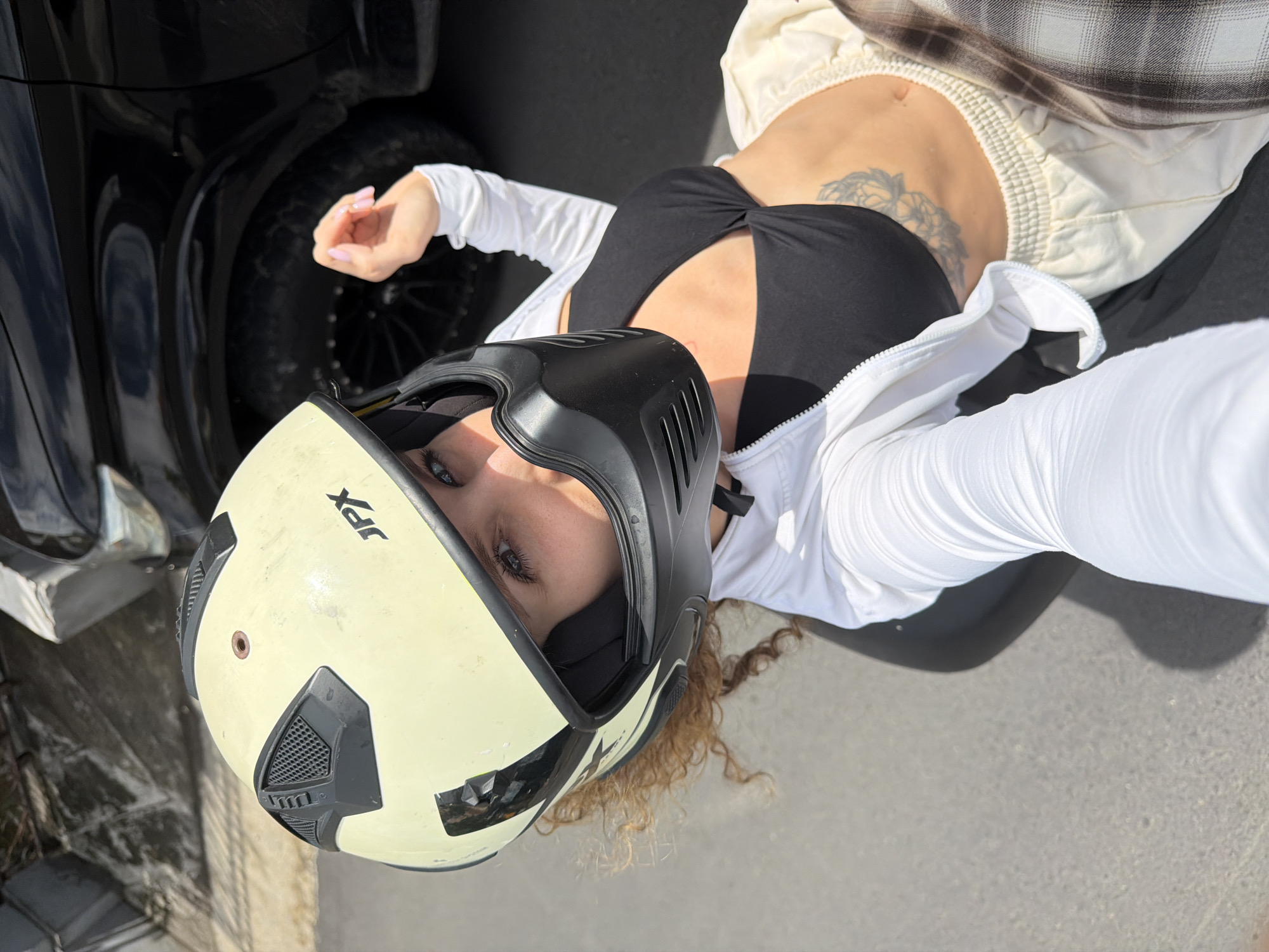 A young woman with curly blonde hair is seated on a motorbike in North Kuta, Indonesia. She is wearing a yellow helmet with a black visor, a black bikini top, a white long-sleeved zip-up shirt, and light-colored sweatpants. Her left hand is raised, with her fingers extended. She is looking towards the camera, with her eyes visible through the helmet visor. Behind her, a black car is parked. The ground is paved. The image appears to be a selfie taken from a high angle.