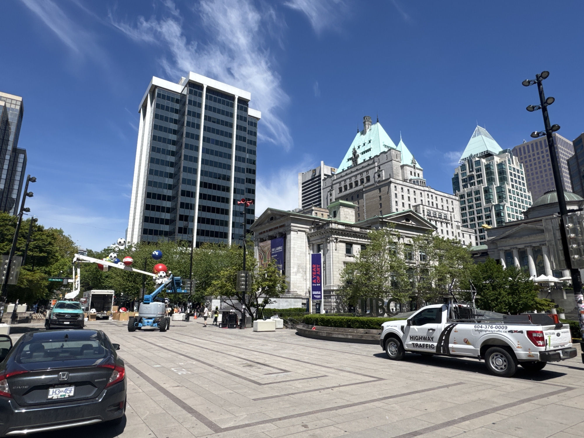 The image shows a city square in Vancouver, Canada, with several notable buildings visible. In the background, there is a tall, rectangular building with a white frame and dark windows. Next to it is a historic-looking building with green turrets. To the right of that, there is another tall, modern building with a similar green tint to its top.
In the foreground, there is a gray Honda Civic with the license plate LH345F parked on the left side of the frame. In front of it, there is a construction or maintenance scene with a white truck, a blue cherry picker, and several large soccer ball decorations being set up. Some people are milling around the area. On the right, there is a white pickup truck with "HIGHWAY TRAFFIC" written on the side, including a phone number (604-376-0009) and an email address. The ground is paved with a pattern of large rectangular stones. In the midground, there's signage for "UBC" and "A YEAR OF ART". The sky is blue with scattered white clouds.