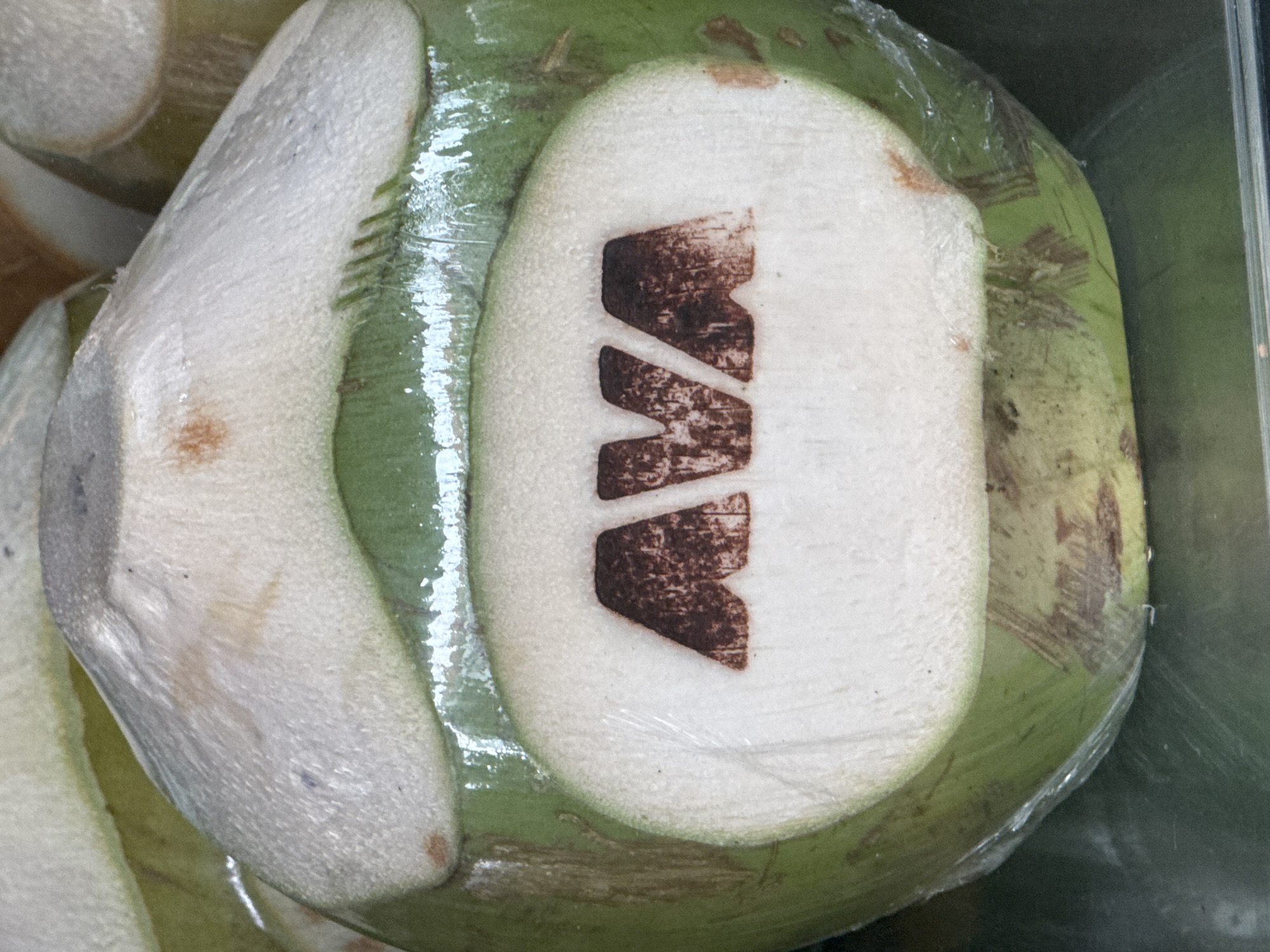 A close-up, eye-level shot shows a fresh green coconut with the letters "AVA" carved into a white, oval-shaped segment on its surface. The coconut is partially wrapped in clear plastic. The carving appears to be done with a dark brown dye or branding. The coconut is resting on a reflective, metallic surface. The background is blurred, showing more coconuts and a warm-toned object, possibly another coconut. The lighting is bright, creating highlights on the plastic wrap and the coconut's surface.