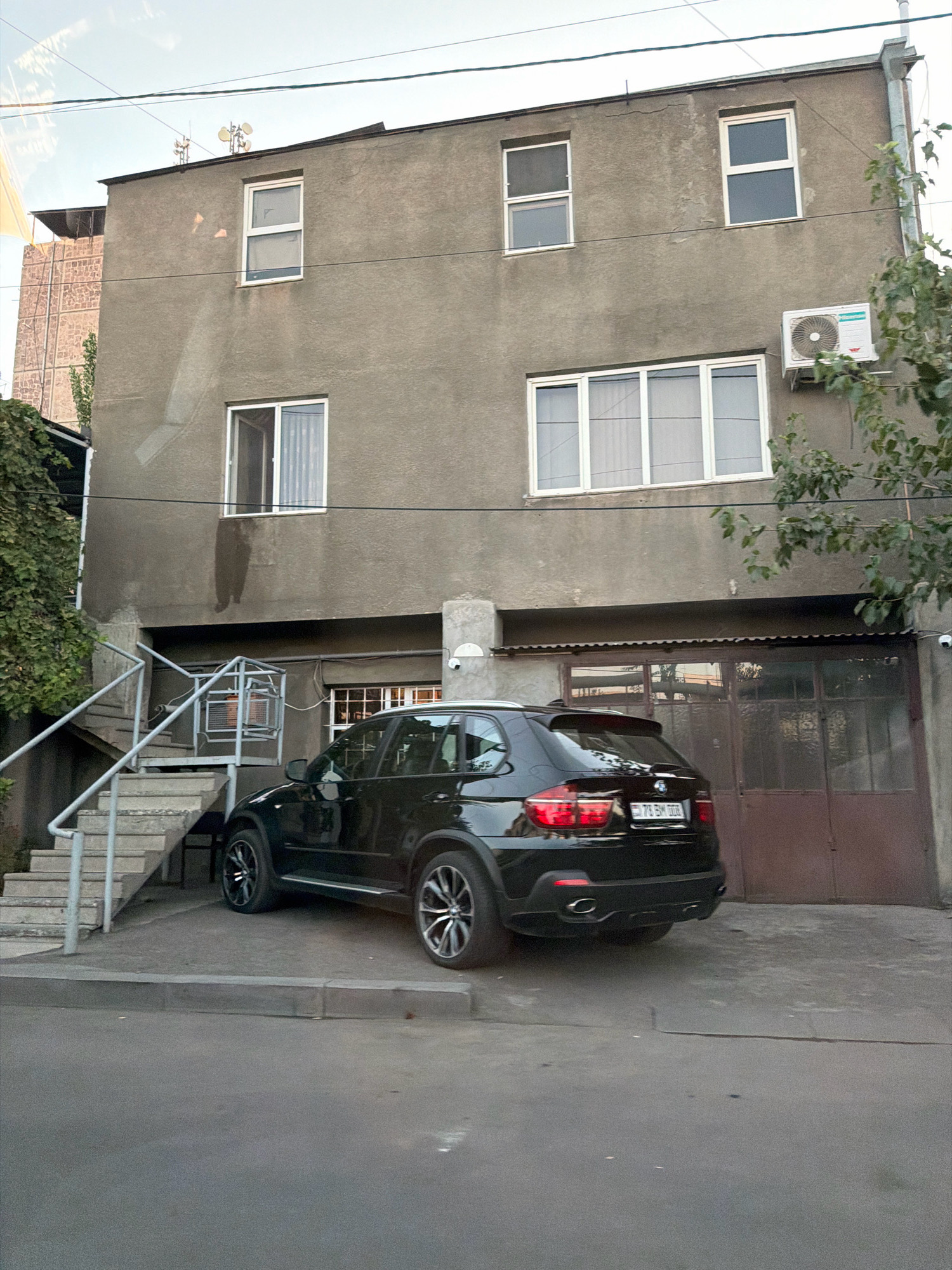 A black BMW X5 SUV is parked in front of a grey, multi-story residential building with multiple windows. An external staircase leads up the left side of the building. The building has a corrugated metal garage door on the ground floor. An air conditioning unit is mounted on the exterior wall. The scene appears to be in a residential area of Yerevan, Armenia, suggested by the license plate on the car.