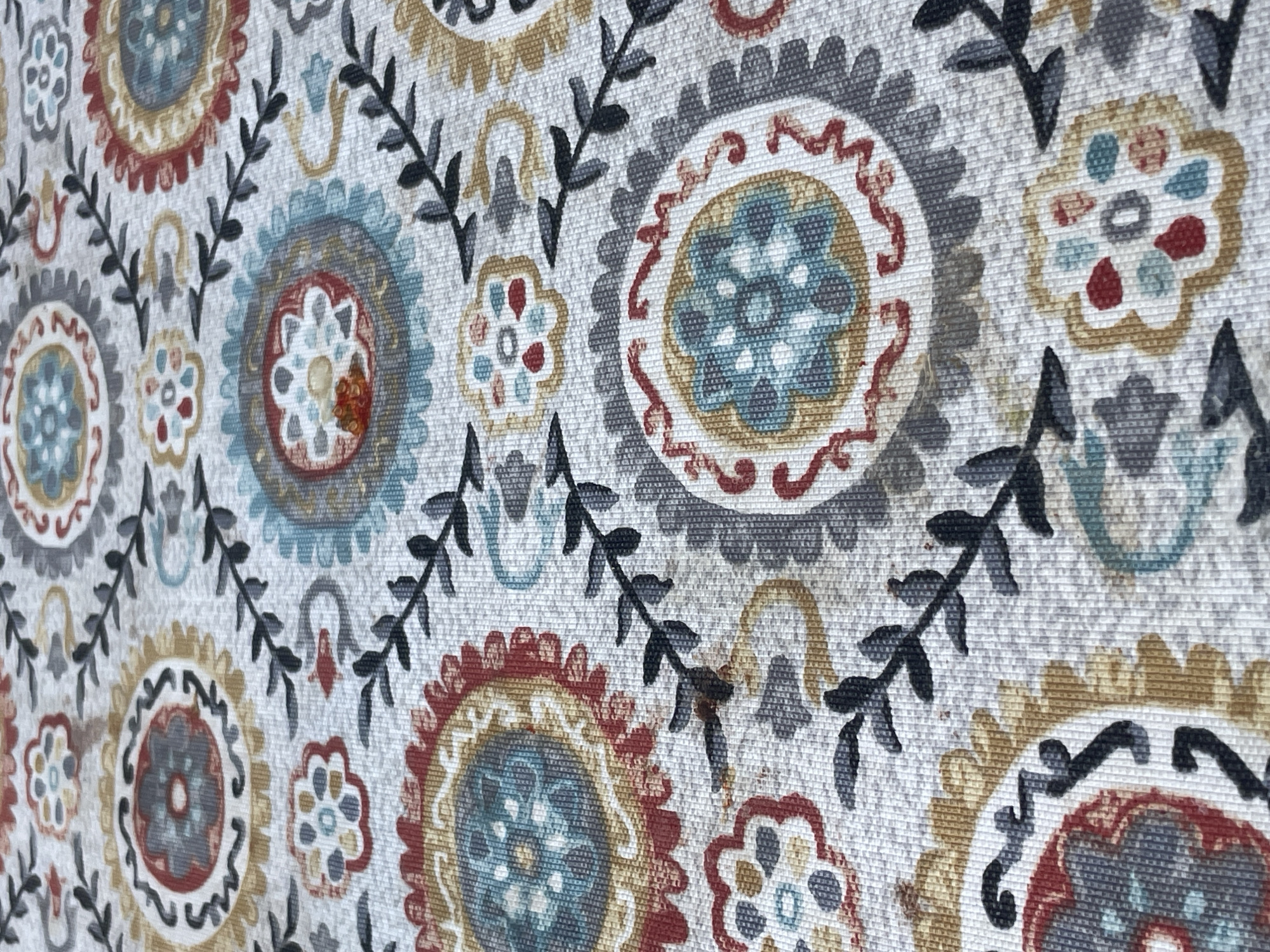 The image displays a close-up, angled view of a patterned fabric. The pattern consists of circular, floral-like motifs in shades of red, blue, yellow, gray, and white, arranged in a repeating tessellation. Interspersed between these larger motifs are smaller, simpler floral shapes and dark gray, stylized vine-like elements with small leaves. The fabric has a textured weave, visible as fine horizontal and vertical lines. There are no people, animals, or discernible elements of an outdoor or urban environment present. This image appears to be of a textile, possibly a tablecloth, rug, or upholstery. Its location in Tashkent, Uzbekistan, cannot be determined from the visual information provided.