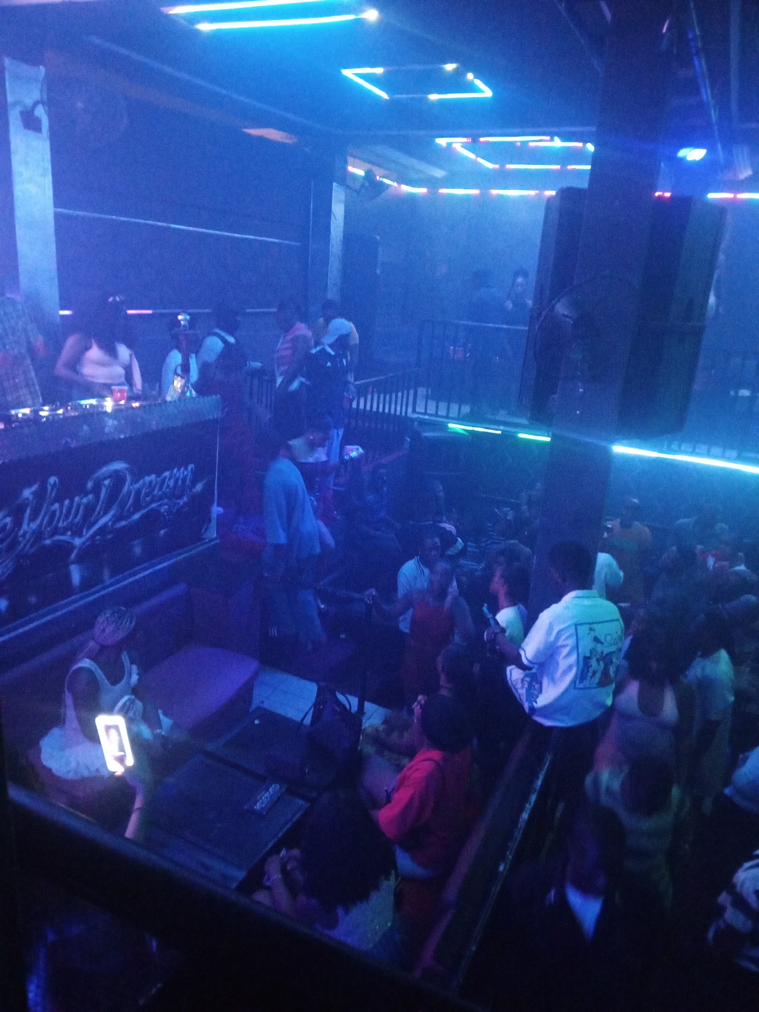 Here is a forensic description of the image:
The image depicts a crowded indoor scene, likely a nightclub or bar, in the city of Unknown, Unknown. The lighting is primarily blue and neon. A DJ booth is visible in the upper left corner. Numerous individuals are present, some standing and interacting, others seated in booths. One person is holding up a cell phone, possibly recording or taking a picture. The environment is dimly lit, obscuring finer details. There is some signage on the wall behind the DJ. Overall, the scene suggests a lively and social atmosphere.