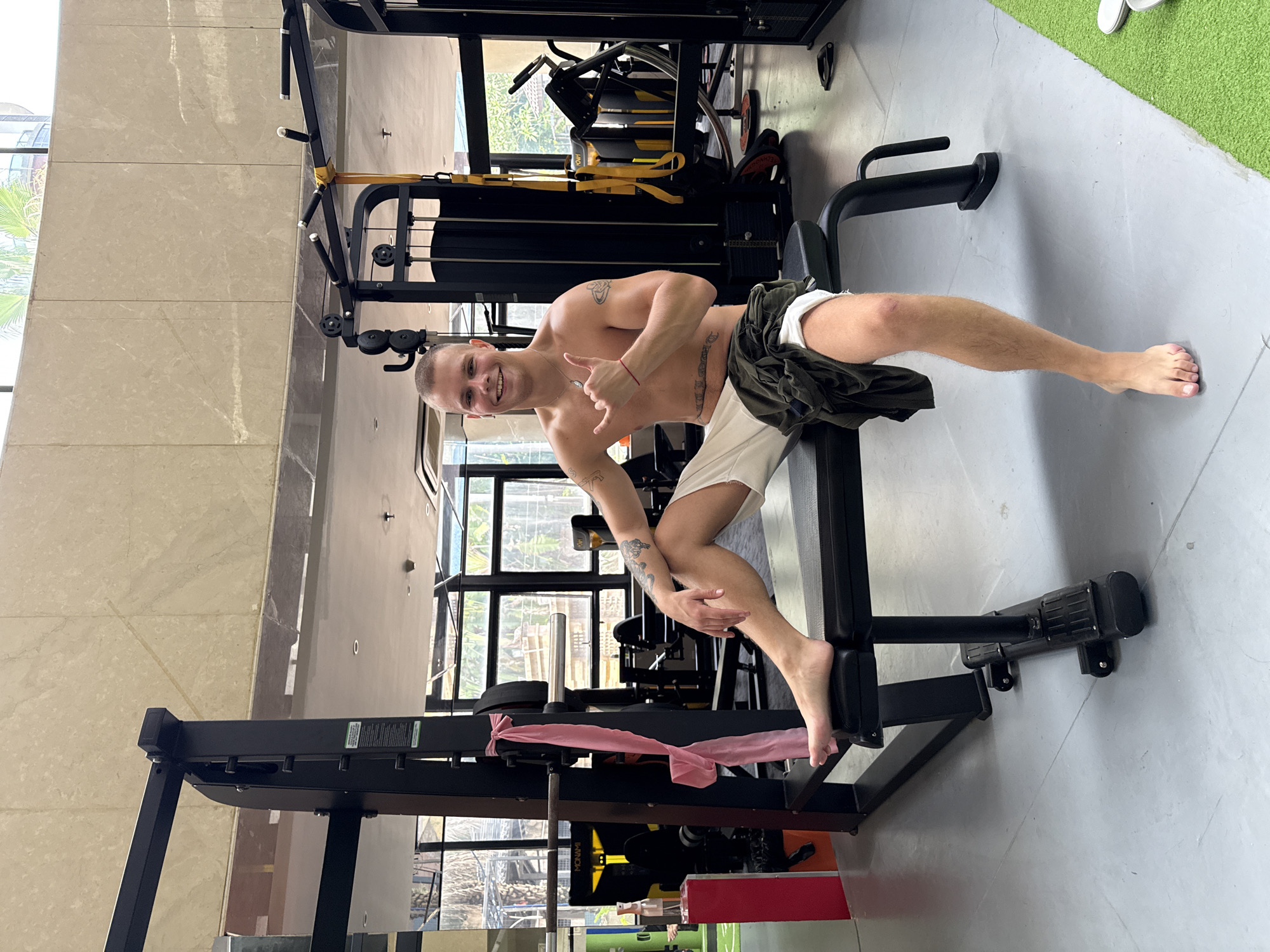 The image depicts a young, light-skinned male sitting on a black exercise bench inside a well-lit gym, likely located in Haizhu District, China. The man has a short buzz cut, is shirtless, and is wearing off-white shorts with a dark green shirt draped over his lap. He is barefoot. He has multiple visible tattoos, including script across his lower abdomen and designs on both upper arms/shoulders. He wears a silver necklace with a pendant and a red string bracelet on his right wrist. He is smiling broadly at the camera, showing his teeth, and his right hand is raised, forming a "shaka" or "hang loose" gesture. His left hand rests on his left thigh.
The gym features light grey concrete flooring, with a section of green artificial turf visible in the lower right corner. The walls are covered in light beige, possibly marble, material. Large, clear windows occupy the upper half of the background, revealing outdoor greenery, palm trees, and parts of other modern buildings. Inside, various black gym equipment is visible. To the left of the man, a black power rack or squat rack is present, with a pink resistance band tied to one of its uprights, and a barbell resting on the j-cups. A red rectangular stand with a clear spray bottle sits next to the rack. Further into the background, a black multi-station cable machine is visible, along with yellow hanging straps (possibly TRX bands) and various black weight plates. A black object with "MONAMI" written in white lettering is on the floor in the background. The ceiling is light-colored and features recessed lighting and smoke detectors.