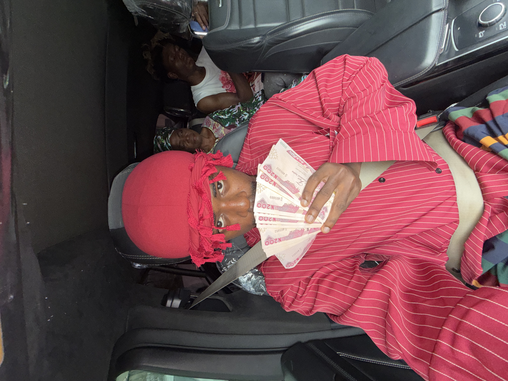 Inside a car in Igboefon, Nigeria, a person wearing a red striped shirt and a red do-rag holds a fan of 200 Nigerian Naira notes. Two other people are lying down in the back of the car. The car has black leather seats.
