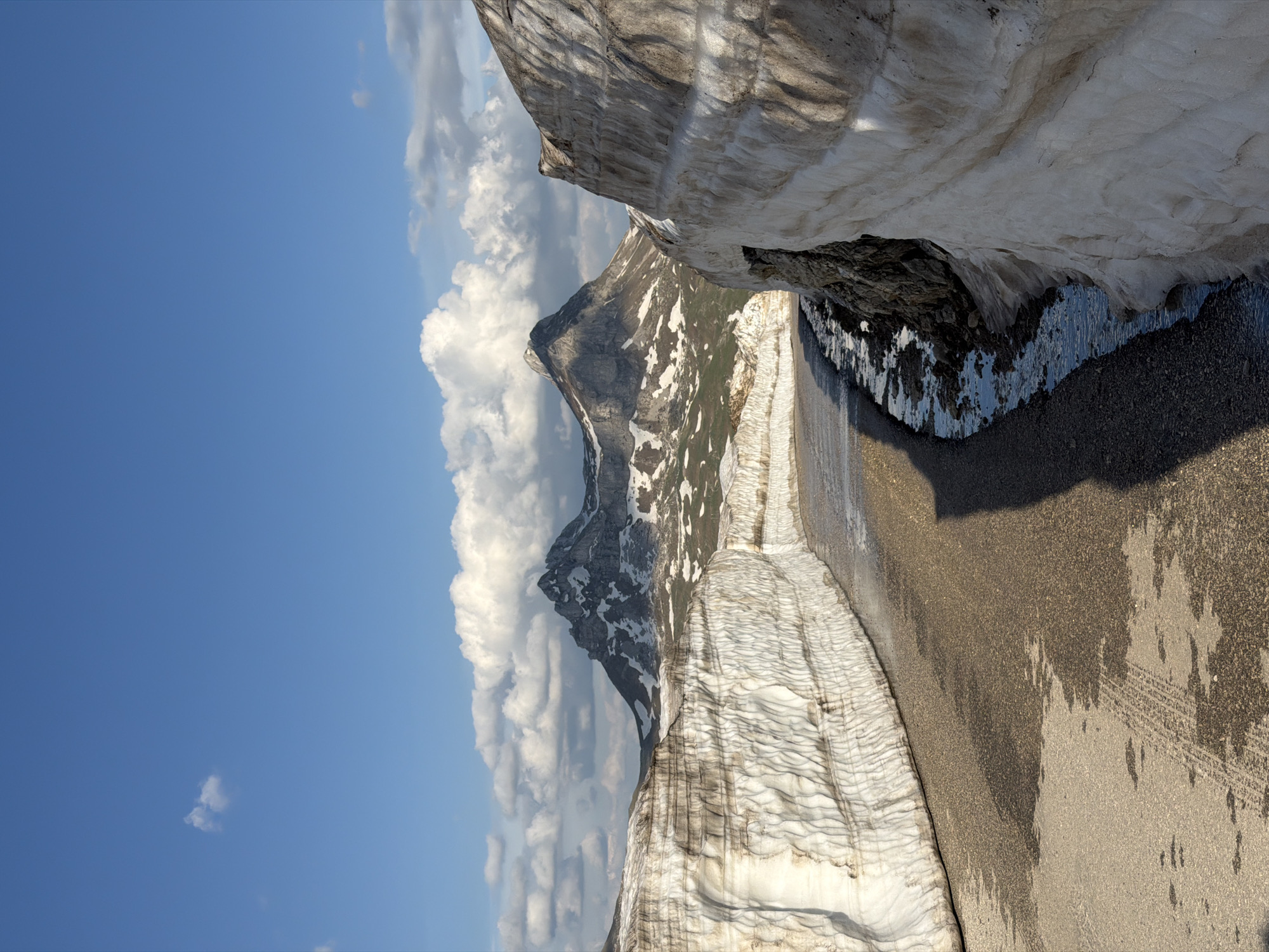 The image shows a mountain landscape under a partly cloudy, blue sky. In the center-left of the frame, a gray mountain peak is visible amidst white clouds. The foreground features a textured surface, with alternating bands of white and brown, possibly representing snow and gravel or dirt. To the right, a darker, uneven surface suggests a road or pathway, with visible tire tracks. The photograph appears to be taken at a high altitude, potentially in a mountainous region, but there is no evidence suggesting the city of Dubrovsko, Montenegro.