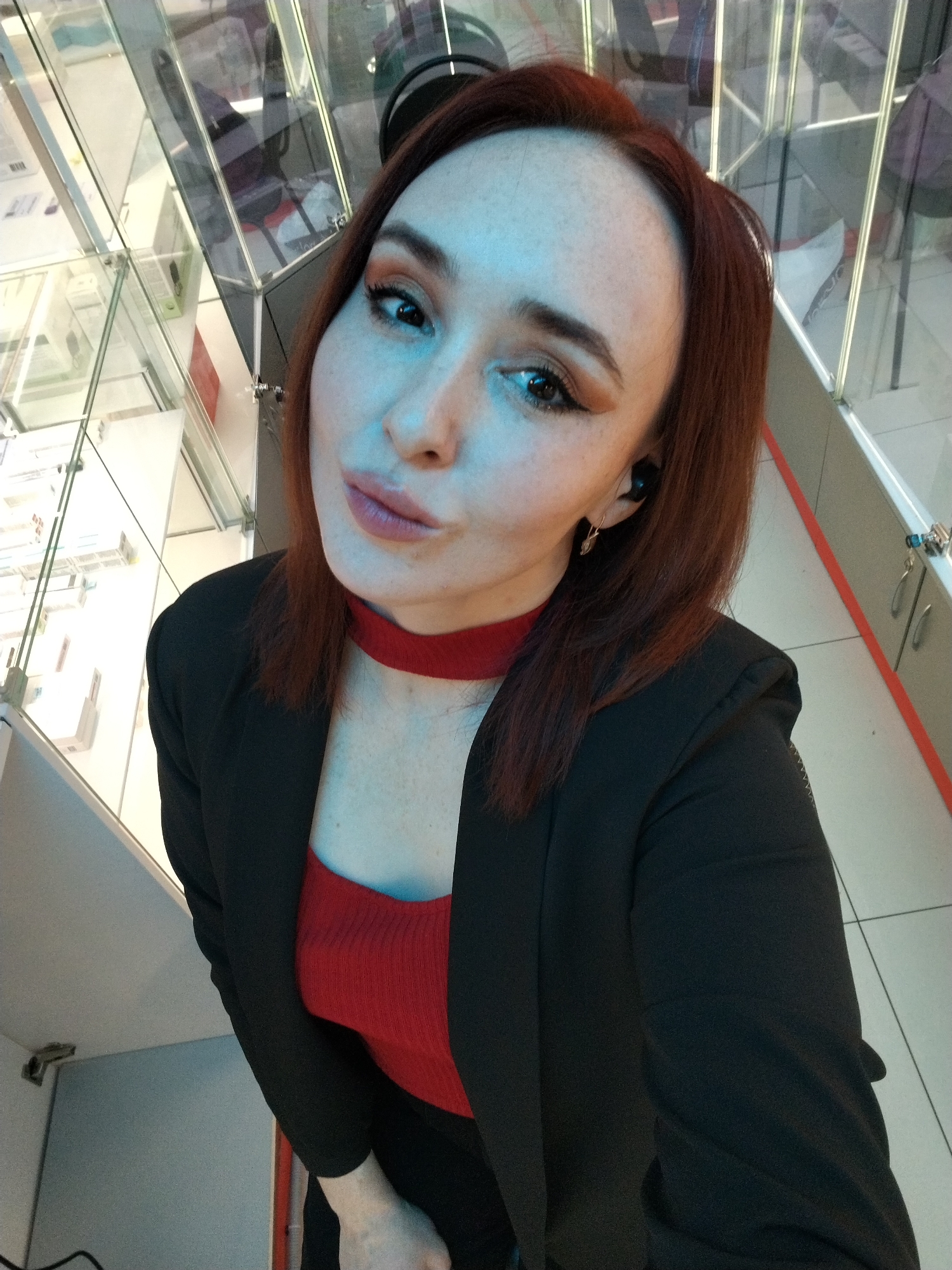A reddish-haired woman wearing a black blazer over a red top and a red choker poses for a selfie in what appears to be a commercial space. The background is partially obscured by glass panels, but some elements suggest a possible store or office environment. Based on the background details and the query, the location could be Izhevsk, Russia.