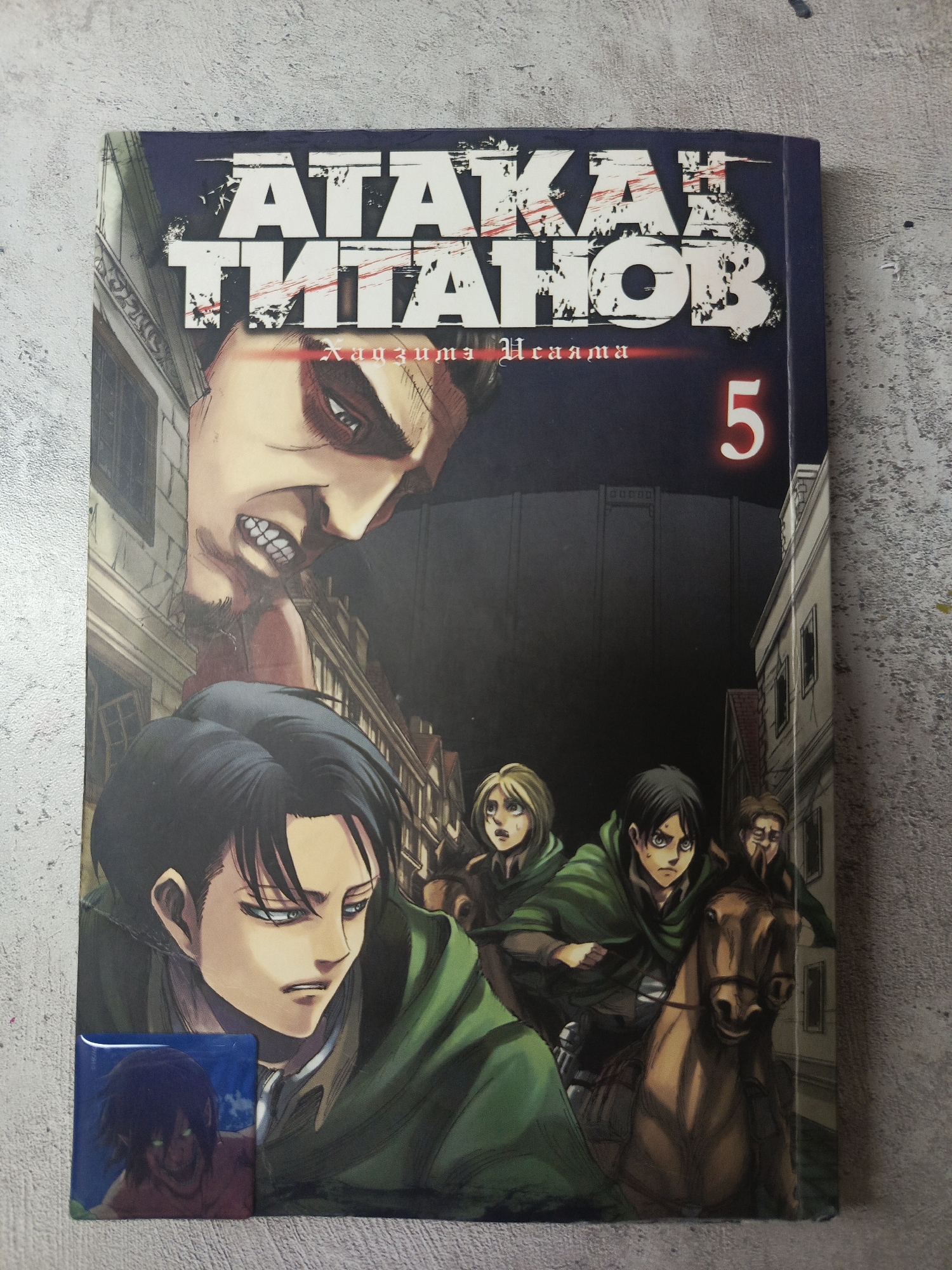The image shows a copy of "Attack on Titan" volume 5. The cover depicts several characters from the manga and anime series "Attack on Titan," including Levi, Eren, and others. A Titan is also prominently featured, looming above the characters. The background shows a cityscape with tall buildings, a wall, and a dark atmosphere. There is a button showing a character (Eren?) in Titan form attached to the book.