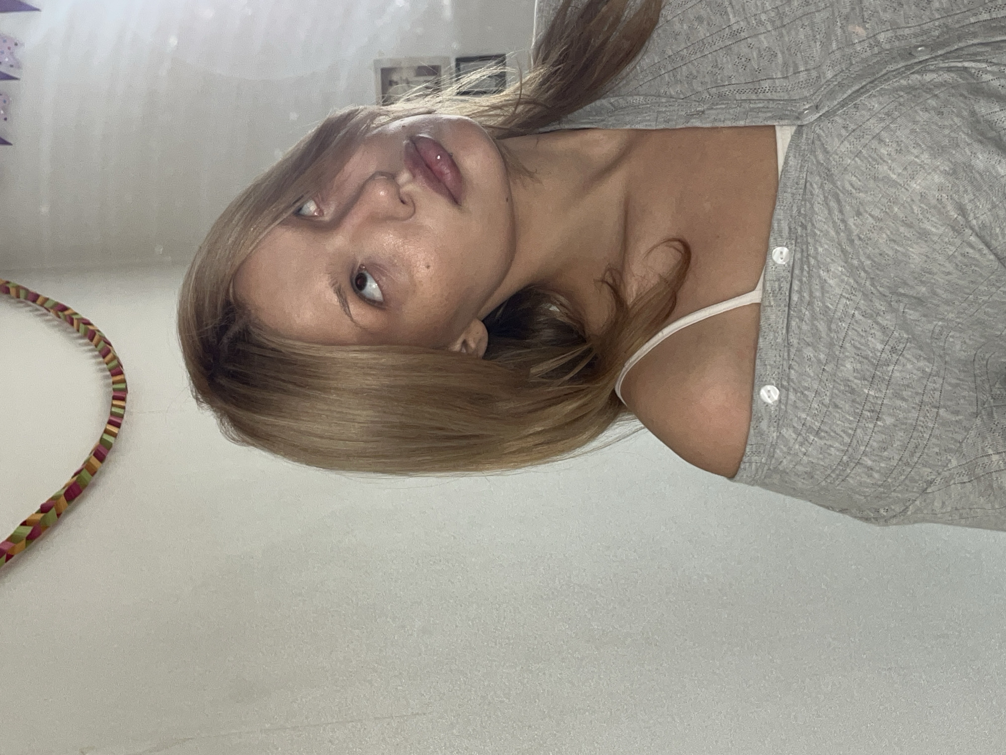 This media file features a young woman captured in a portrait shot, oriented sideways with her head on the left and body extending to the right. She has light brown hair, fair skin, and appears to be looking upwards and slightly to her right (relative to her head position). Her lips are slightly parted, and there's a small glint on her upper lip, possibly a reflection or a subtle piercing. She wears a light grey, textured or ribbed, long-sleeved top or cardigan, unbuttoned, over a white camisole or bralette with thin straps.
The background is a simple, light-colored wall, suggesting an indoor setting, likely a room. In the upper right corner (if the image were upright), some blurred framed pictures or decorations are visible on the wall. A colorful, braided or wrapped object, possibly a hula hoop with green, red, and yellow stripes, is partially visible in the lower left corner.
The lighting is soft and diffuse, suggesting natural light, possibly daytime. The woman's expression is neutral, appearing calm or contemplative as she poses for the photograph. There is no discernible text in the image. The location context provided is Chaikovsky, Russia.