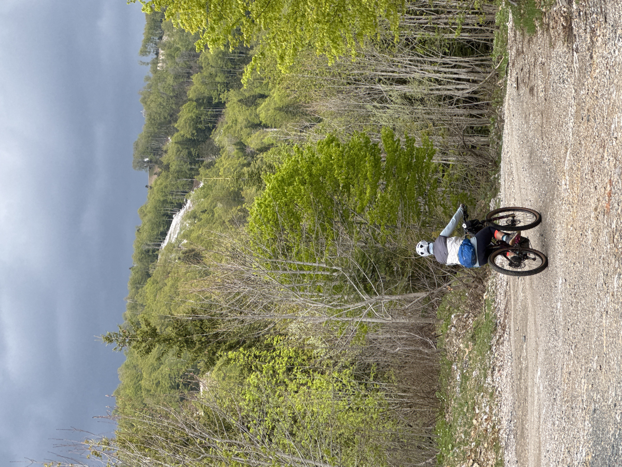 A person on a mountain bike rides along a gravel road in a mountainous environment. The person is wearing a helmet, light-colored long-sleeved shirt, dark pants, and colorful socks. They have a small blue bag attached to their back. The road is gravelly and slopes downward. The surrounding environment is heavily forested with deciduous trees, many just beginning to leaf out. A section of snowy terrain appears on a distant hillside. The sky is overcast, with a dark, ominous cloud formation present. The location is Katun Bačko brdo, Montenegro.