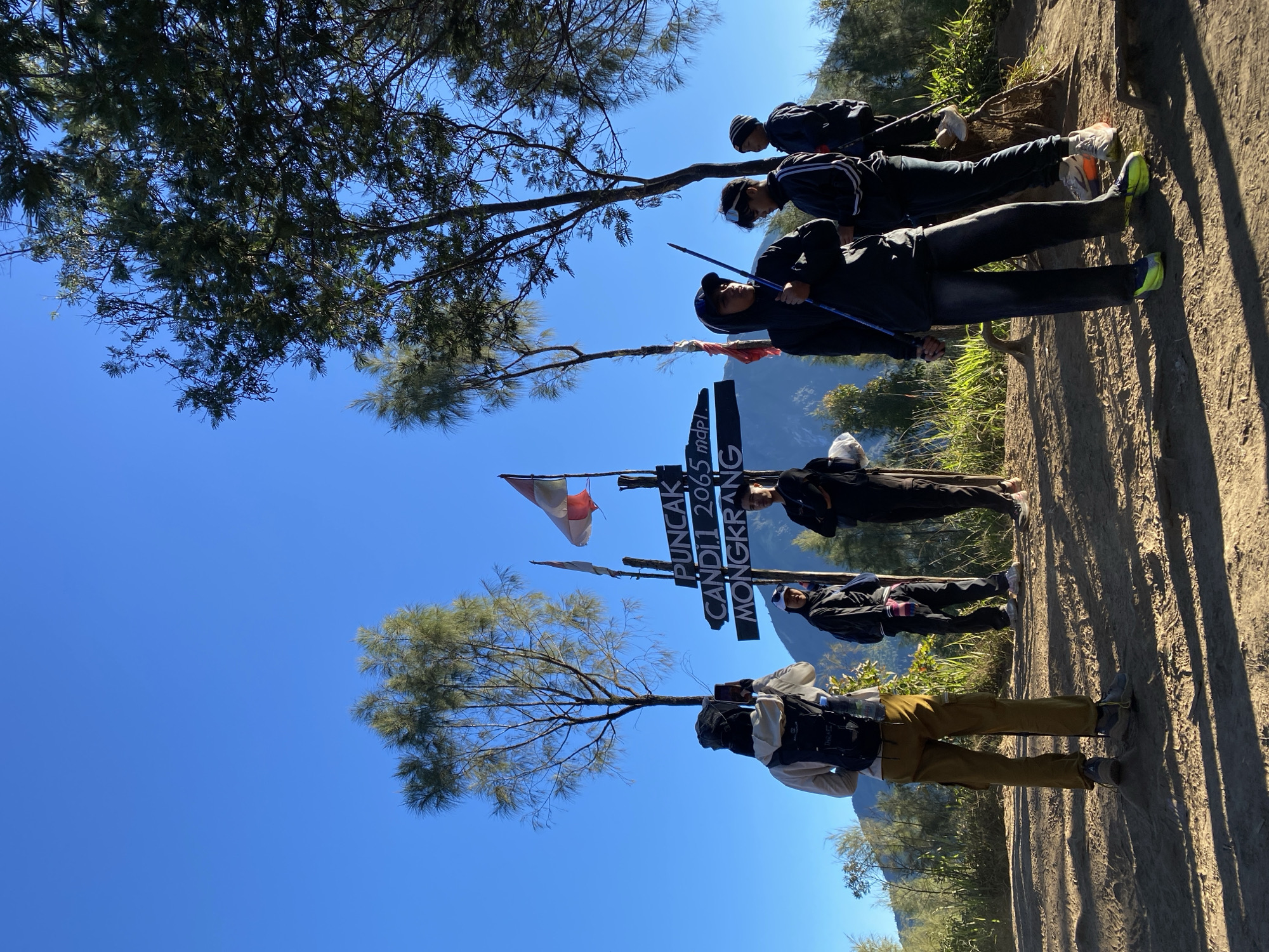 In Gondosuli, Indonesia, a group of six people stands near a signpost on a sunny day. The signpost, adorned with flags, indicates "PUNCAK CANDI 2065 mdpl MONGKRANG." One person in the foreground is taking a photo with a smartphone, while others stand around the signpost, some holding hiking poles. They are dressed in casual outdoor attire, including backpacks and hats, suggesting a hiking or trekking activity. The environment is natural, with sparse trees, dry ground, and visible shadows cast by the sun. In the background, mountains are partially visible under a clear blue sky.