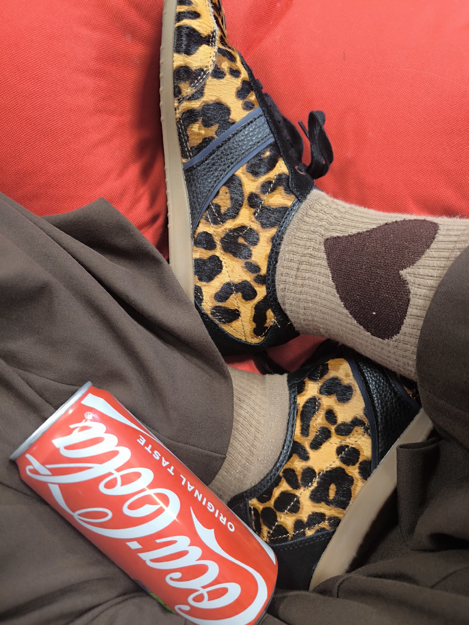 The image shows a person wearing leopard-print sneakers and socks with a brown heart, seated on a red cushion. A can of Coca-Cola is positioned on the brown fabric near the person's legs. The specific location in Riga, Latvia is not identifiable from the image.