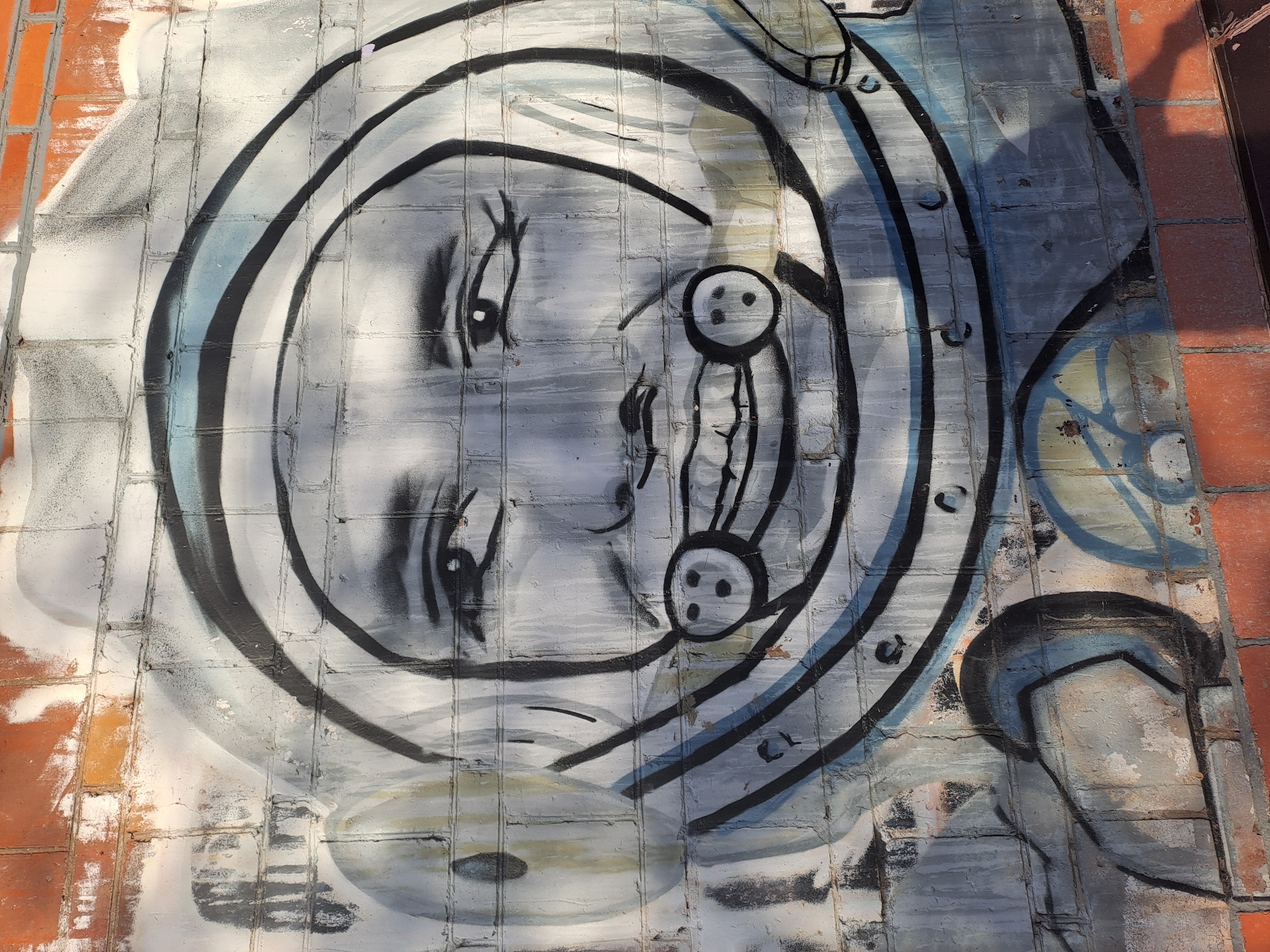 The image shows a piece of street art, likely graffiti, on a tiled surface in Elektrostal, Russia. The artwork depicts a stylized human face inside a circular shape, resembling a diving helmet or porthole. The face is pale with simplified features, large eyes, and a geometric representation of a nose and mouth. The art is drawn with thick black lines on a white or light grey background, and the tiles are a mix of light grey and brick-red colors. The sunlight casts a shadow on the upper right.