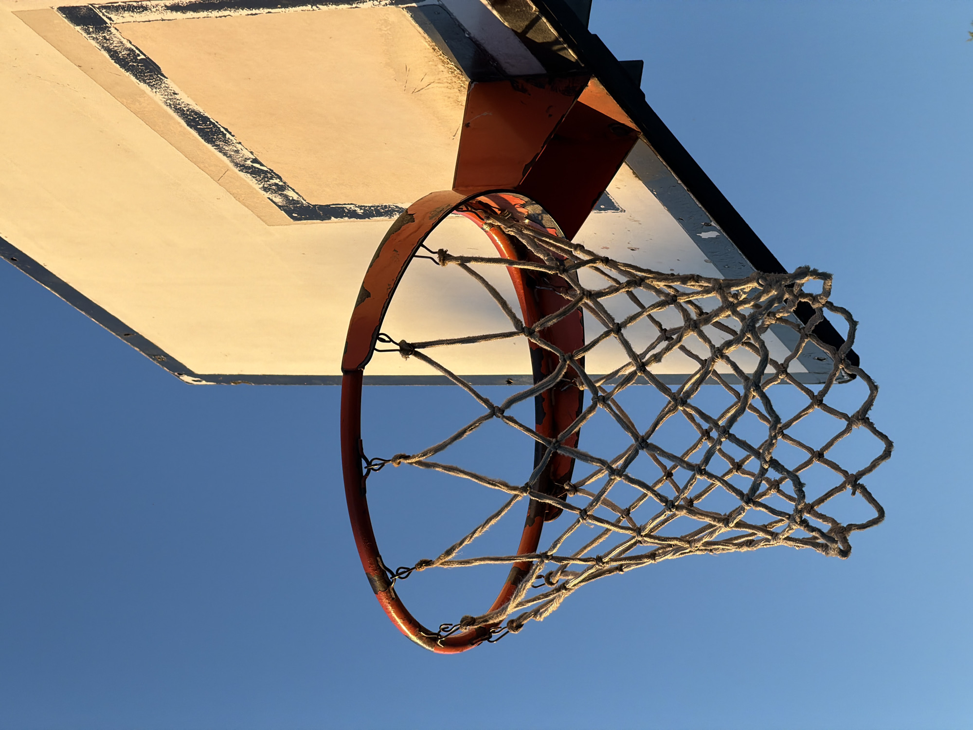 A worm's-eye view captures a basketball hoop and backboard against a clear blue sky. The backboard is rectangular, with a faded white surface and worn edges marked by dark paint. The hoop, painted orange and showing signs of rust and wear, is attached to the backboard via a metal support structure. A net of woven white rope hangs below the hoop. The scene suggests an outdoor basketball court, potentially in a public park or schoolyard. There is no evidence of this image being taken in Milan, Italy.