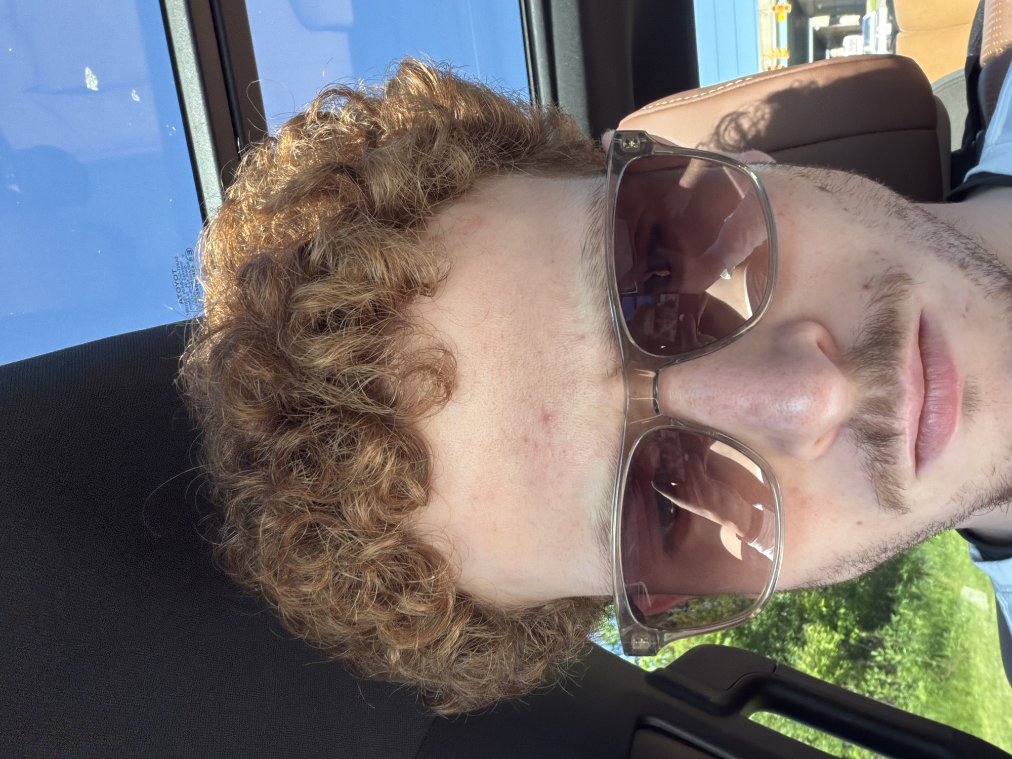 A close-up, rotated image captures a person's head and upper torso inside a vehicle. The subject has light skin, prominent curly light brown hair, and light facial stubble around the chin and upper lip. They are wearing sunglasses with clear, somewhat thick frames and dark, reflective lenses that obscure their eyes. The reflections in the sunglasses show bright sunlight and distorted elements of the car's interior and exterior.
The setting is clearly inside a car, with a black textured surface visible in the lower left, likely a seat or dashboard. Behind the subject, a brown leather headrest or seat upholstery is visible. Through the car window, a vibrant blue sky and lush green trees or foliage are seen, indicating an outdoor, natural environment. Bright sunlight illuminates the scene, suggesting a clear, sunny daytime.
On the car window, text is visible, including a reversed "ATOYOT" which is likely the "TOYOTA" logo, along with "E3" and other alphanumeric markings, confirming the vehicle's manufacturer. The image appears to be a close-up selfie or taken by a passenger, focusing primarily on the person's face. The person's expression is neutral and largely concealed by the sunglasses. The location context is Pepper Pike, United States of America.