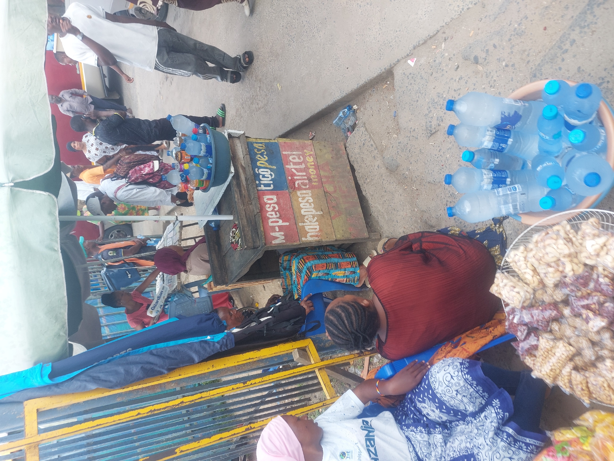 The image captures a bustling scene in Dar es Salaam, Tanzania. Several individuals are gathered, some seated on blue plastic chairs near a blue and yellow metal barrier. A woman in a red dress and another in a white top with blue patterns and a pink headscarf are prominent. A wooden stand displays signs for "M-pesa Tigo Pesa" and "Hale Pesa Airtel Money," indicating mobile money services. A bucket filled with bottled water and a basket of peanuts are visible, suggesting vendors selling these items. Other individuals stand nearby, their attire varying from white t-shirts to patterned clothing. The overall environment appears to be a busy street or marketplace.