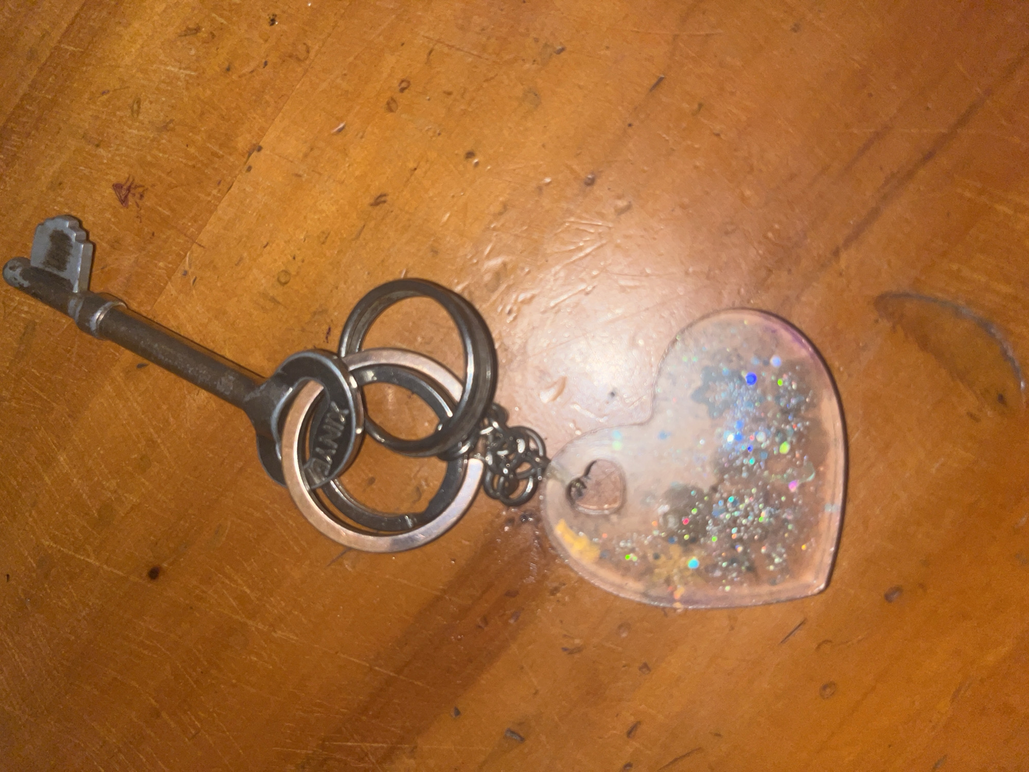 An overhead close-up shot captures a vintage-style metal key and an attached heart-shaped keychain resting on a wooden surface. The surface is a warm, orange-brown hue with visible wood grain, scratches, and minor imperfections, suggesting an old table or floor.
The key is made of dark, aged metal with an ornate, possibly skeleton-key style, handle and a long shaft. It is connected to several keyrings. One large keyring is silver-toned, another appears to be copper or rose gold-colored, and a smaller dark metal ring links them to the heart-shaped charm. Faint, partially legible text, possibly "VINTAGE," is visible on the copper-colored keyring.
The keychain itself is a clear, translucent heart, likely made of resin or plastic, filled with a vibrant mix of glitter in various colors including silver, blue, pink, green, and gold. A small, solid copper-colored heart charm is embedded within the clear heart. The objects are static, with no activity taking place, under what appears to be indoor lighting.
