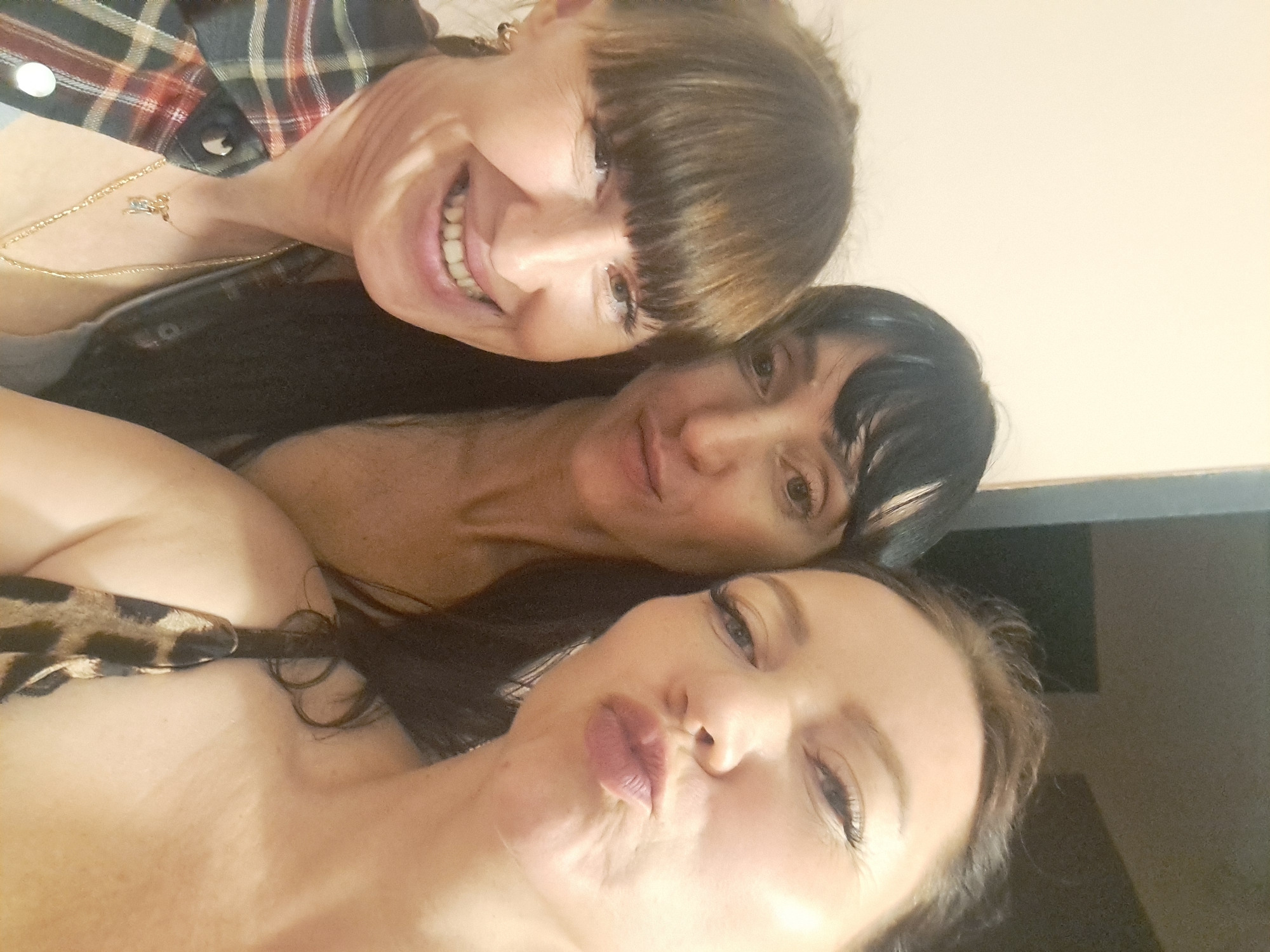 Three women are captured in a close-up selfie against a plain, cream-colored background. The woman on the left is smiling broadly, her head tilted slightly. She has brown hair with bangs and is wearing a plaid shirt. The woman in the middle has dark hair and is looking towards the camera with a neutral expression. The woman on the right is puckering her lips as if for a kiss, her face angled towards the camera. She has light brown hair and is wearing a leopard-print top. The lighting is somewhat dim, with a slight glare on the faces.