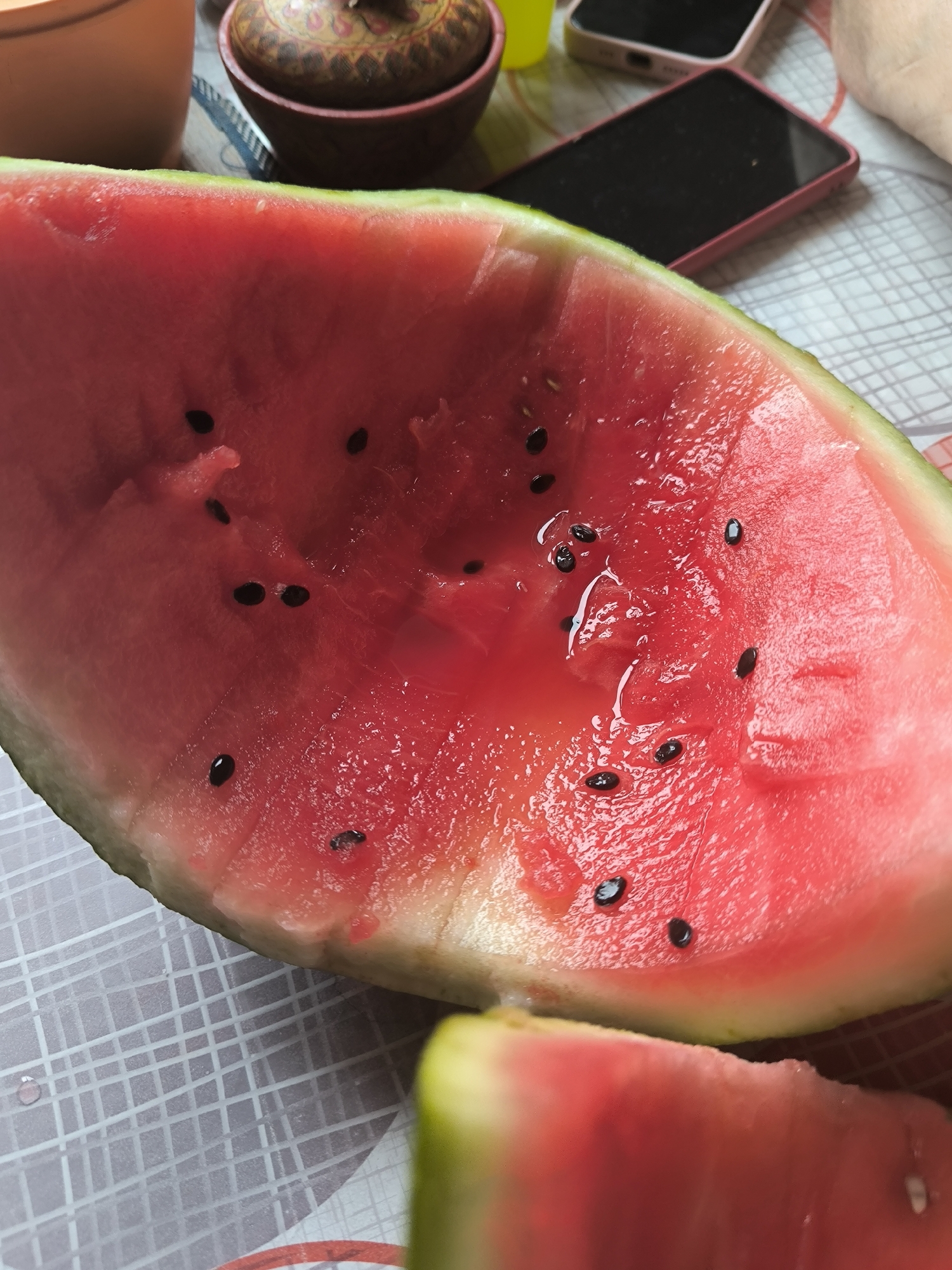 A close-up, overhead shot displays a slice of ripe watermelon, its vibrant pink flesh studded with black seeds, resting on a white and gray checkered surface. To the left, a portion of a beige ceramic container is visible, and behind it, a decorative wooden bowl with intricate carvings. In the background, two smartphones are laid out, one with a dark screen and the other with a pink casing. A small yellow cup also appears in the background. The scene is indoors, likely on a table. No specific location in Maiskii, Russia, can be determined from the visual information provided.