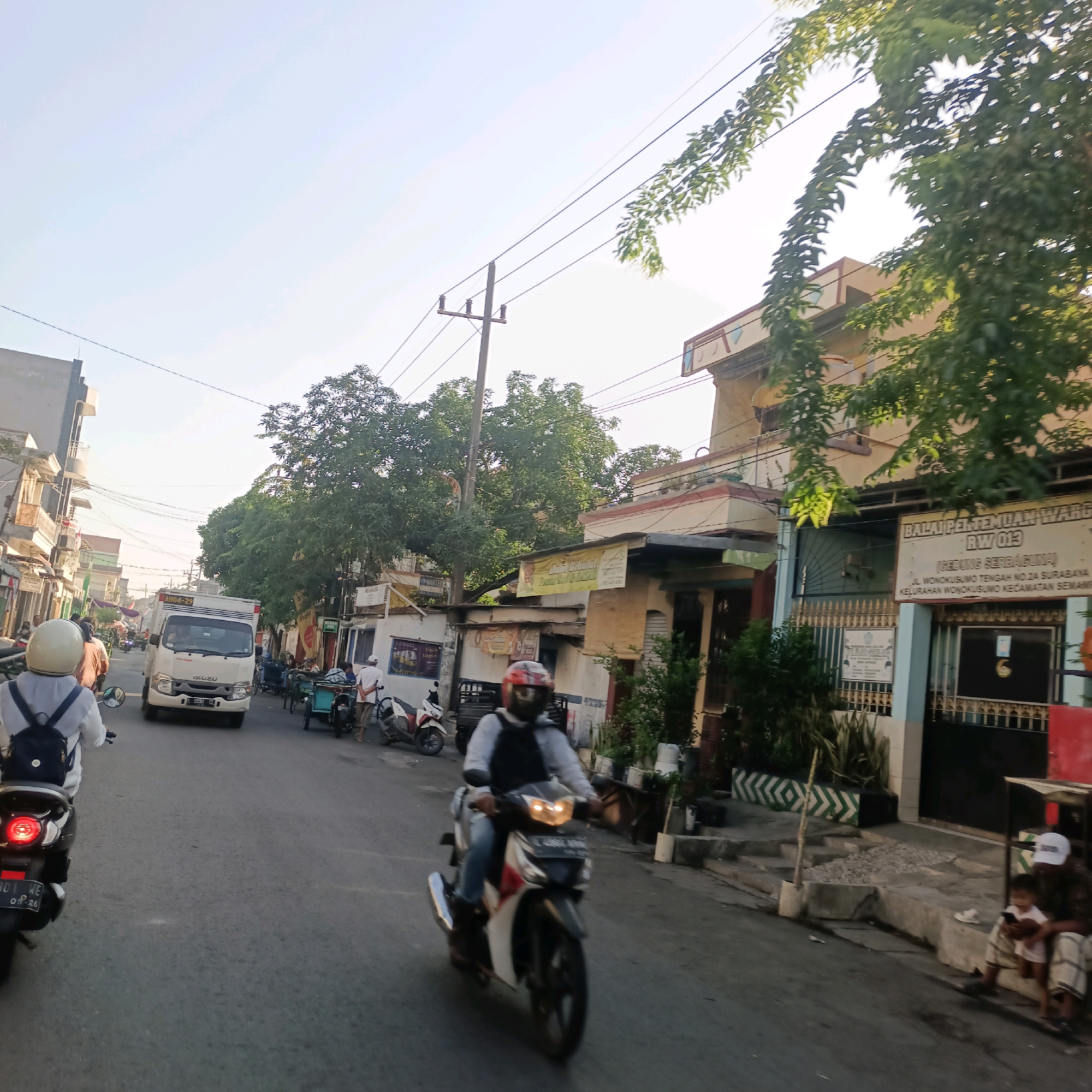 The image depicts a street scene in Surabaya, Indonesia, likely in a residential or commercial area. The time of day appears to be late afternoon, with sunlight casting long shadows and illuminating the sky with a pale blue hue. The street is paved and shows signs of traffic, with several vehicles visible.
In the foreground, a person on a motorcycle is riding away from the camera. The motorcycle is dark-colored with a red taillight, and the rider is wearing a white shirt and a backpack. Further down the street, another motorcycle rider, wearing a red and white helmet, is approaching.
A white Isuzu truck is positioned in the middle of the road, appearing to be in motion or stopped. On the right side of the street, there are buildings with signs, one of which reads "BALAI PERTEMUAN WARGA RW 013" and "JL. WONOKUSUMO TENGAH NO.2A SURABAYA." This indicates the location is the RW 013 (Neighborhood Community) Meeting Hall on Wonokusumo Tengah Street No. 2A in Surabaya.
The buildings along the street have various architectural styles, with some featuring tiled roofs and decorative elements. Trees with lush green foliage are interspersed throughout the scene, providing shade and adding to the urban landscape.
On the sidewalk to the right, a person in a white cap is sitting next to a small child, who appears to be looking at a mobile phone. Other individuals and various objects like bicycles and carts are also present, suggesting a typical street life in a populated area. The overall impression is a snapshot of everyday life in a busy Surabaya neighborhood.