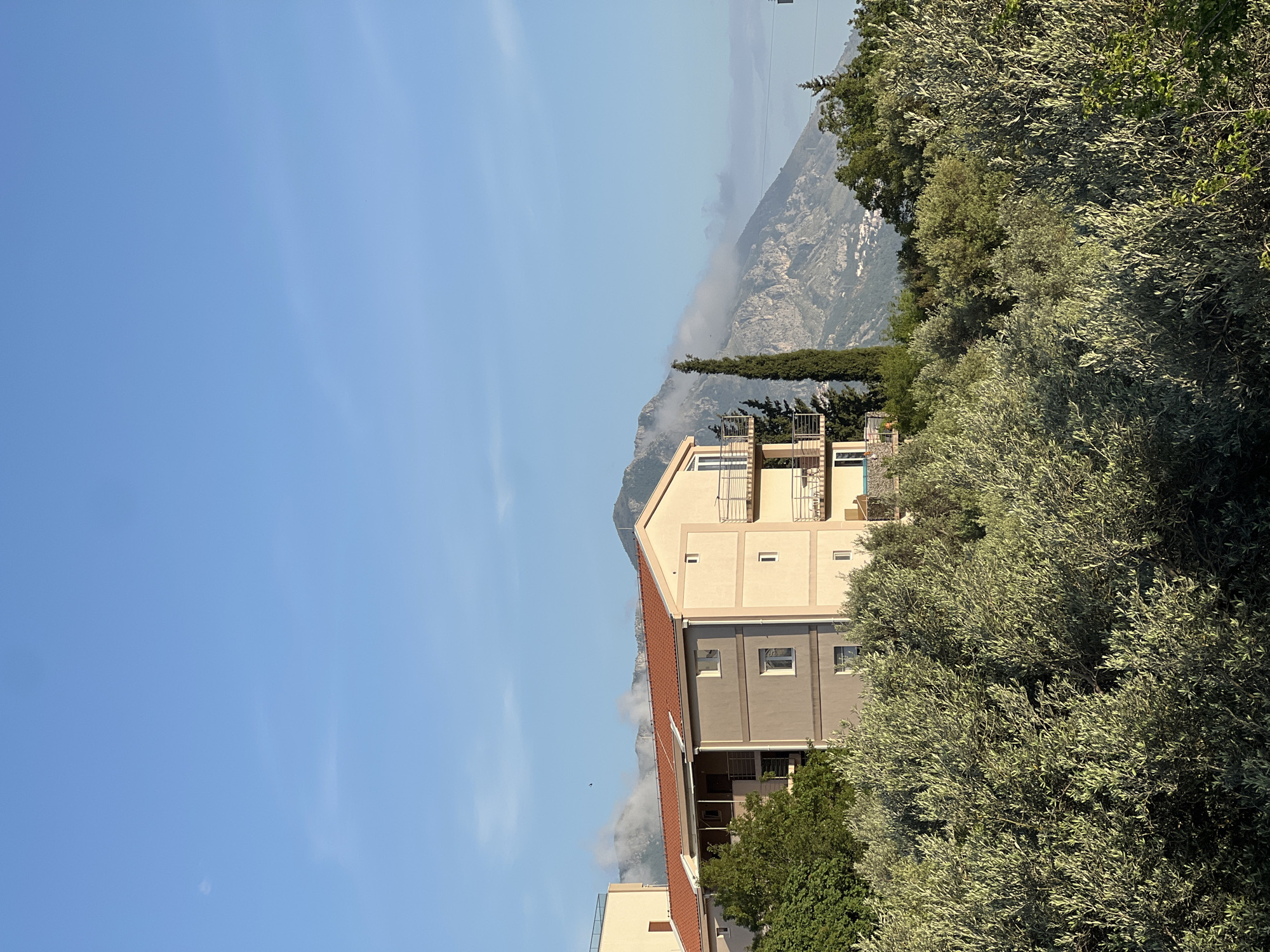 A medium shot shows a building in Ivanovići, Montenegro, with a mountain in the background. The building has an orange-tiled roof and beige and gray sides. There are green trees in front of the building. The sky is blue with light clouds. A mountain range is behind the building, partially obscured by clouds.
