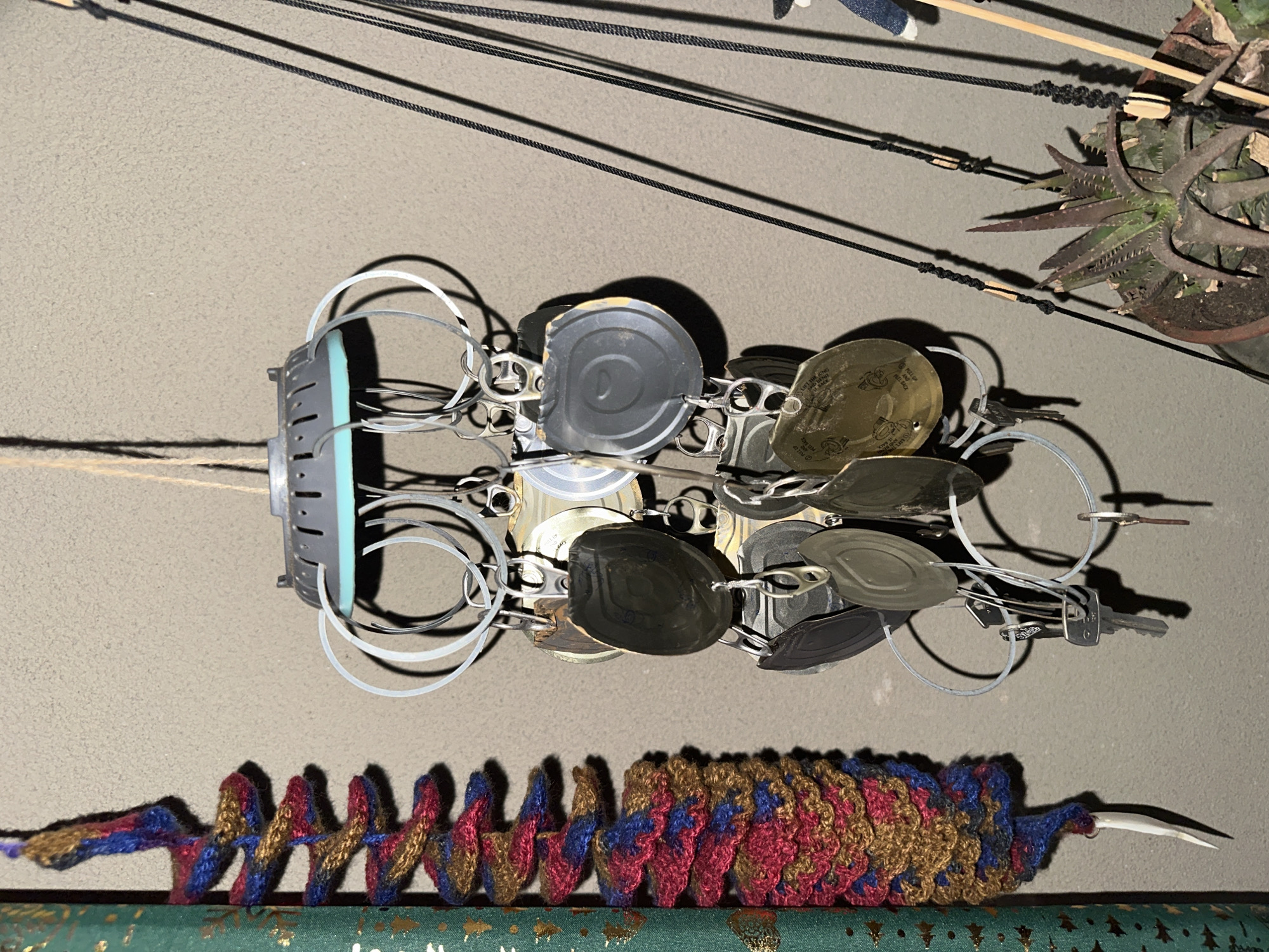 The image depicts an assemblage of objects arranged against a neutral-toned wall, indoors, and its location in Arica, Chile, is not visually discernible.
Dominating the center is a constructed object that appears to be a kinetic sculpture or mobile. It features a gray-and-light-blue central component from which multiple metal rings are suspended. Attached to these rings are various metal objects, primarily can lids, and possibly keys and other small metallic elements.
Below this mobile is a colorful, knitted, worm-shaped object with red, blue, gold and brown hues. It rests on a surface covered in green wrapping paper with golden patterns.
Above the mobile, thin black strings or wires are strung horizontally across the frame. To the upper right, a potted succulent plant is partially visible.
The lighting is somewhat diffused and casting soft shadows, indicating an ambient or artificial light source. There are no people depicted in the image.