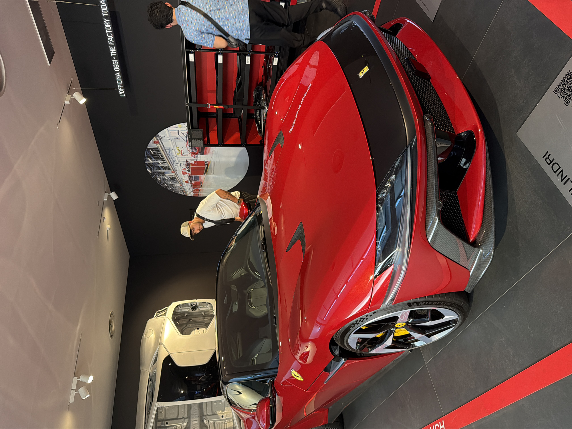 In Maranello, Italy, at the Ferrari museum, a bright red Ferrari SF90 Stradale is showcased on a polished dark grey floor. The car's interior features grey leather seats. Two men are present; one, wearing a white t-shirt and a baseball cap, stands near the car, looking at it. The second man, in a blue patterned shirt and black pants, stands further away, facing a black shelving unit filled with car parts. The wall behind the car displays a large, circular mural of a factory interior and a neon sign that reads "L'OFFICINA OGGI - THE FACTORY TODAY." The overall environment suggests a modern exhibition or showroom dedicated to Ferrari's manufacturing and history.