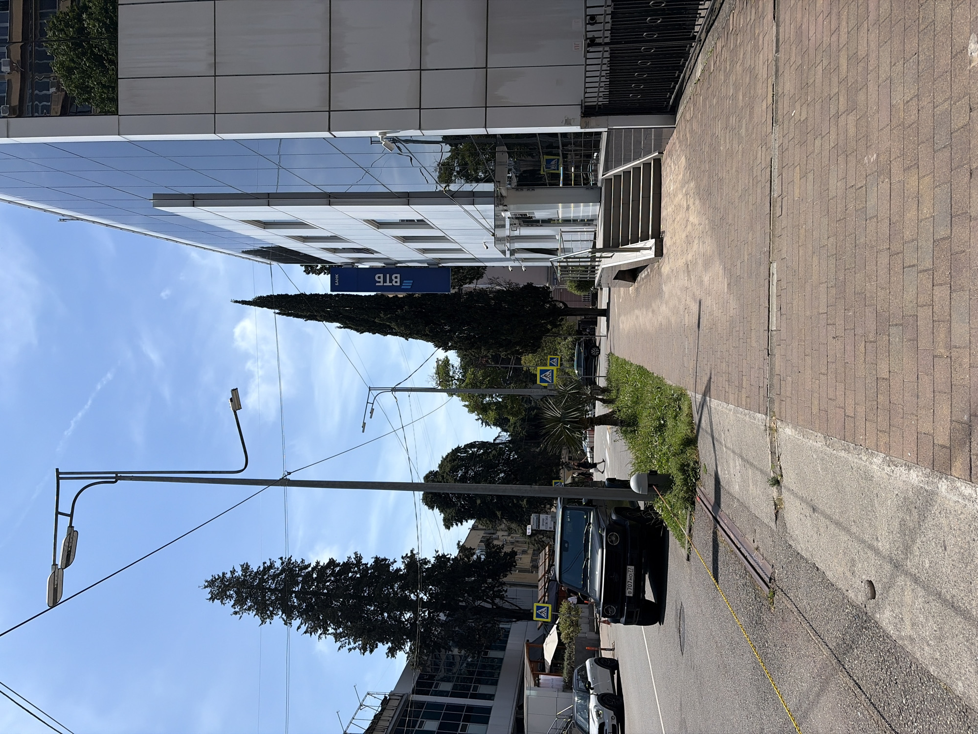 This image depicts a sunny, clear daytime street scene at Ulitsa Ostrovskogo, 41, Sochi, Krasnodar Krai, Russia.
On the right side of the frame, a modern, multi-story building with a reflective glass facade and white paneling is prominently featured. A vertical blue sign on the building displays "ВТБ" and "БАНК" in white Cyrillic text. An entrance is accessible via a set of light-colored stairs, approximately six steps, equipped with a metal handrail. A dark metal fence borders the property line on the far right.
To the left of the building, an asphalt road runs parallel, marked with a yellow line. A black Mercedes-Benz G-Wagen, bearing Russian license plate "В 707 НО 77," is parked partially on the asphalt and a narrow grassy strip. A white Mini Cooper is partially visible further down the road on the left. The street is lined with tall, dark evergreen trees and smaller, palm-like trees. Several yellow diamond-shaped signs with black pedestrian symbols are positioned along the street.
The foreground is a sidewalk paved with brown rectangular bricks, separated from the road by a concrete curb. A light grey cylindrical bollard is present on the grassy strip adjacent to the parked G-Wagen. In the midground, a tall grey street light pole with multiple light fixtures extends over the road. Numerous black overhead wires are visible against the clear blue sky, which contains a few wispy white clouds and a faint contrail in the upper left.
Several indistinct pedestrians are visible walking in the midground, both on the main sidewalk and further down the street. The scene is brightly illuminated by direct sunlight, casting clear shadows on the ground and structures.