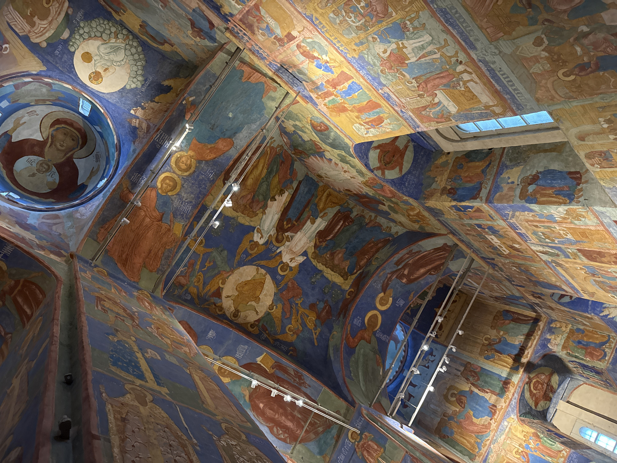 The image depicts the interior of the Church of the Nativity of the Theotokos in Suzdal, Russia, showcasing its extensive frescoed walls and ceilings. The artwork features religious scenes with numerous figures rendered in vibrant blues, reds, oranges, and yellows against a blue background. The frescoes cover the vaulted ceilings and walls, creating a densely decorated and immersive environment. Several figures, including angelic beings and human individuals in historical attire, are depicted. The scene includes architectural elements such as arches and a window with a blue-tinted pane. Lighting fixtures are visible on the ceiling. The perspective is looking upwards, emphasizing the grandeur and intricate detail of the painted interiors.