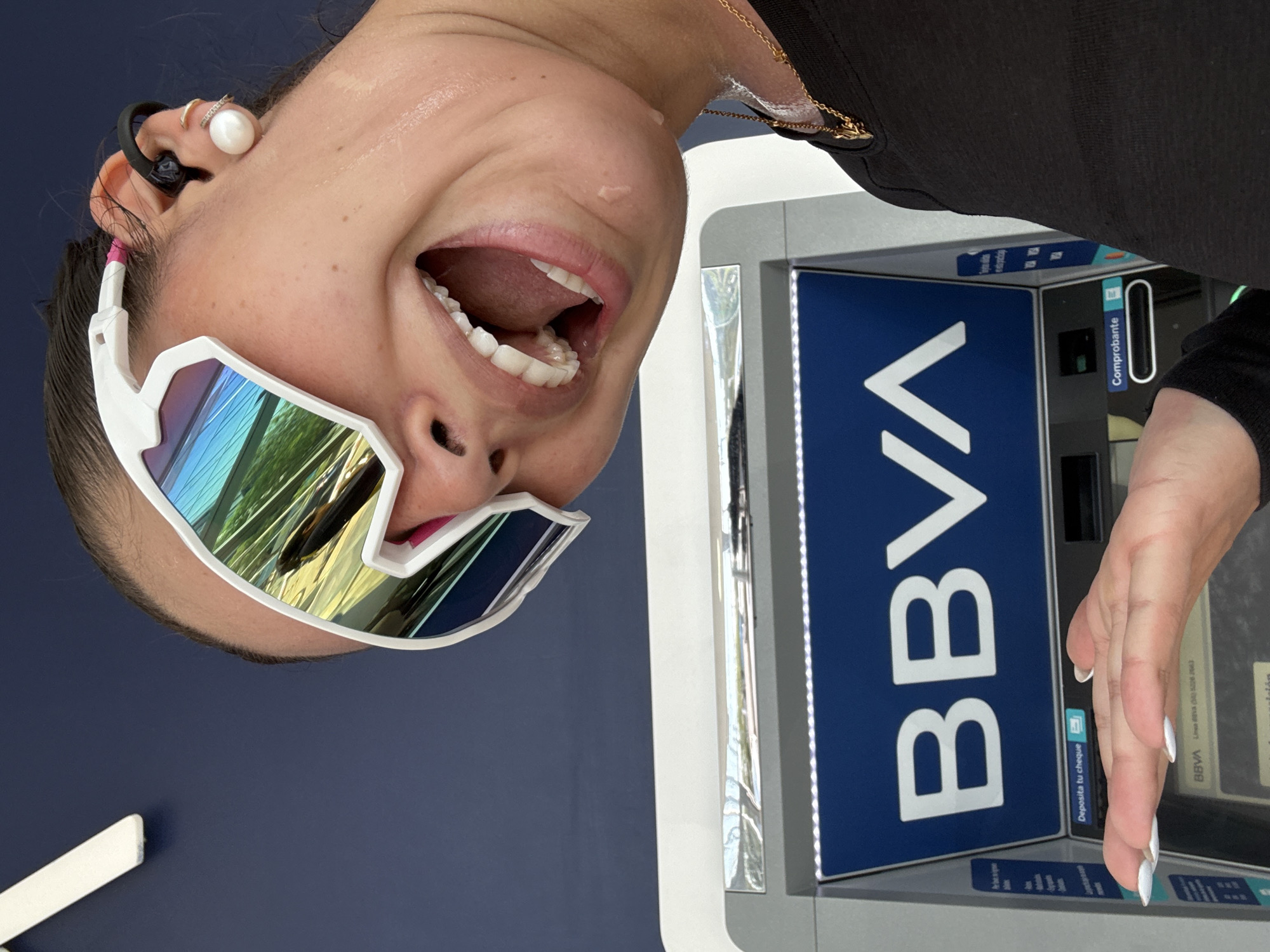 A woman wearing white sunglasses with reflective lenses, a black top, and a pearl earring with a ring is standing in front of a BBVA ATM. The ATM is blue and white. The woman has her mouth open and appears to be excited. The ATM is located in Puebla, Mexico.