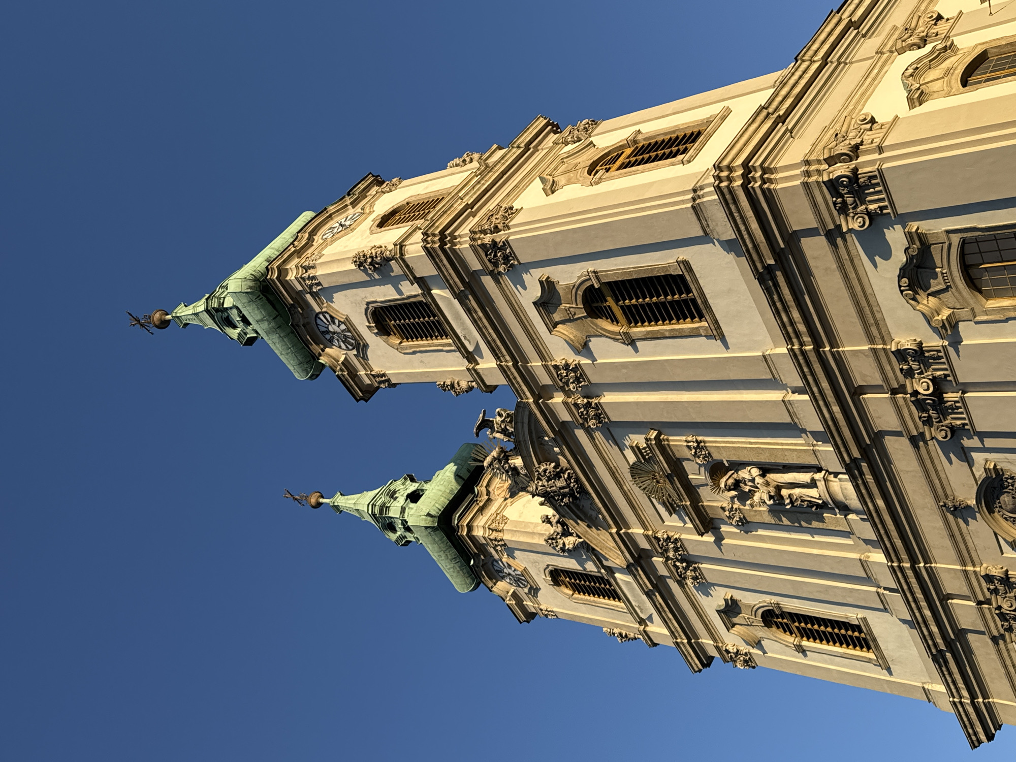 The image displays a grand, ornate Baroque-style building, likely a church or cathedral, viewed from a low angle against a clear, deep blue sky. The building features two prominent towers with distinctive patinated green, spire-like roofs and finials at their peaks. The main facade of the building is a light, possibly off-white or cream color, richly adorned with darker gray or beige architectural details such as pilasters, cornices, decorative window surrounds, and intricate carvings. Several windows with grates are visible along the building's sides.
The sunlight, with a golden hue, strongly illuminates one side of the structure, casting the other side into shadow and creating pronounced contrasts that highlight the three-dimensional relief of the architecture. This lighting suggests either early morning or late afternoon during the "golden hour." Clocks are visible on at least one of the towers. Intricate sculptures and decorative elements, including a statue of a figure within a niche, are visible on the facade. The overall impression is one of grandeur and detailed craftsmanship. No discernible text is visible. Given the provided context, this elaborate architectural structure is located in Budapest, Hungary.