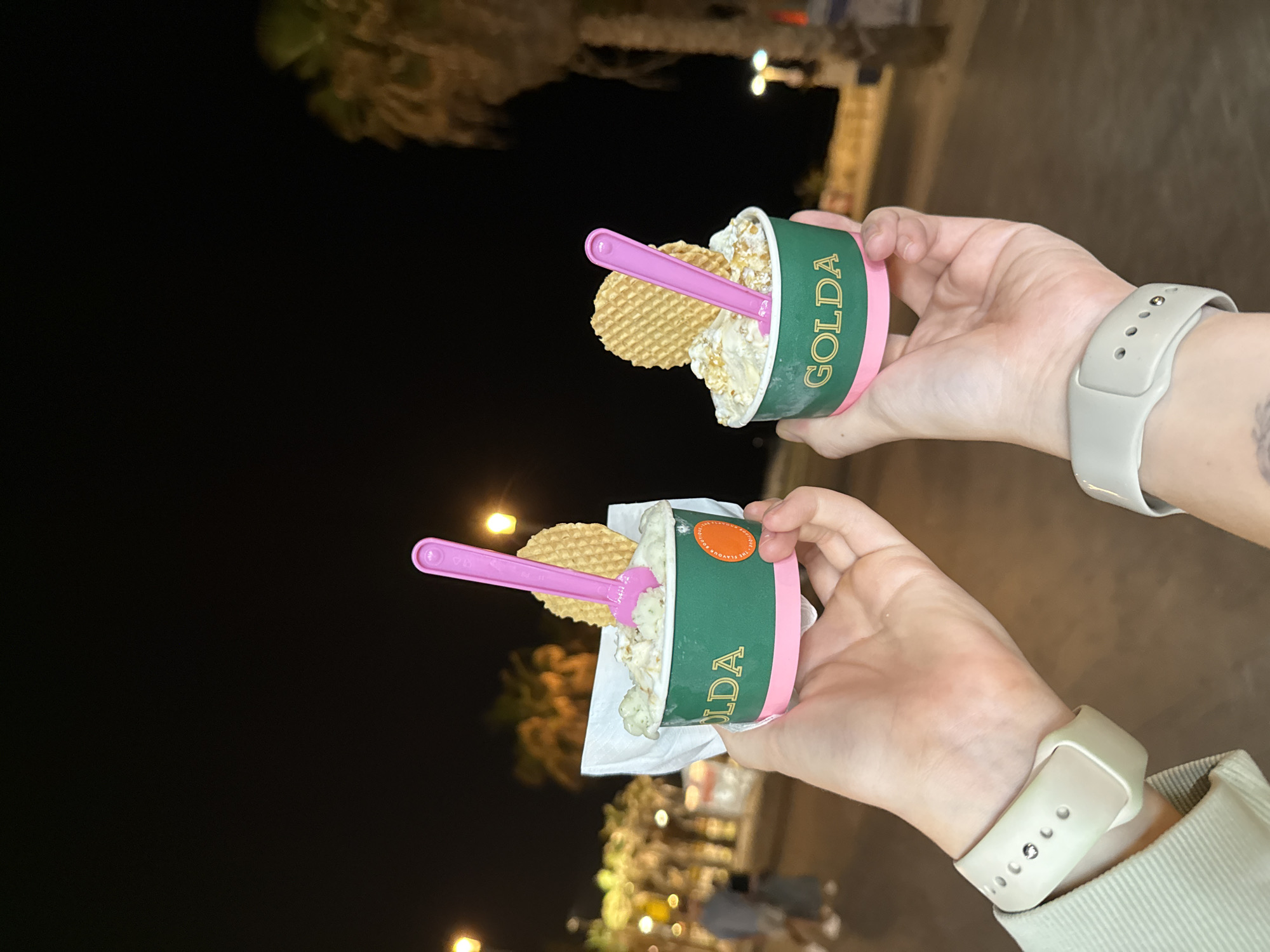 A nighttime outdoor photograph, rotated 90 degrees counter-clockwise, depicts two hands holding cups of "Golda" gelato. The dark background is softly illuminated by blurred, warm-toned artificial lights, suggesting an urban or promenade setting in the evening, consistent with Tel-Aviv, Israel.
Both hands hold identical "Golda" gelato cups, which feature a dark green upper band with "GOLDA" printed in gold letters, and a pink base. Each cup contains a light-colored gelato, possibly with visible toppings like nuts or crumbled biscuits. A small, round, waffle-textured cookie and a pink plastic spoon are inserted into the gelato of each cup. On the bottom cup, a white paper napkin is wrapped around its base, and part of a circular logo on the cup is visible, with faint text including "THE FLAVOUR."
The hand at the top of the frame wears a light grey silicone smartwatch, with a tattoo partially visible on the wrist. The hand at the bottom wears a light beige silicone smartwatch, and the sleeve of a light-colored jacket or shirt is visible. The image captures a casual moment of enjoying ice cream outdoors at night.