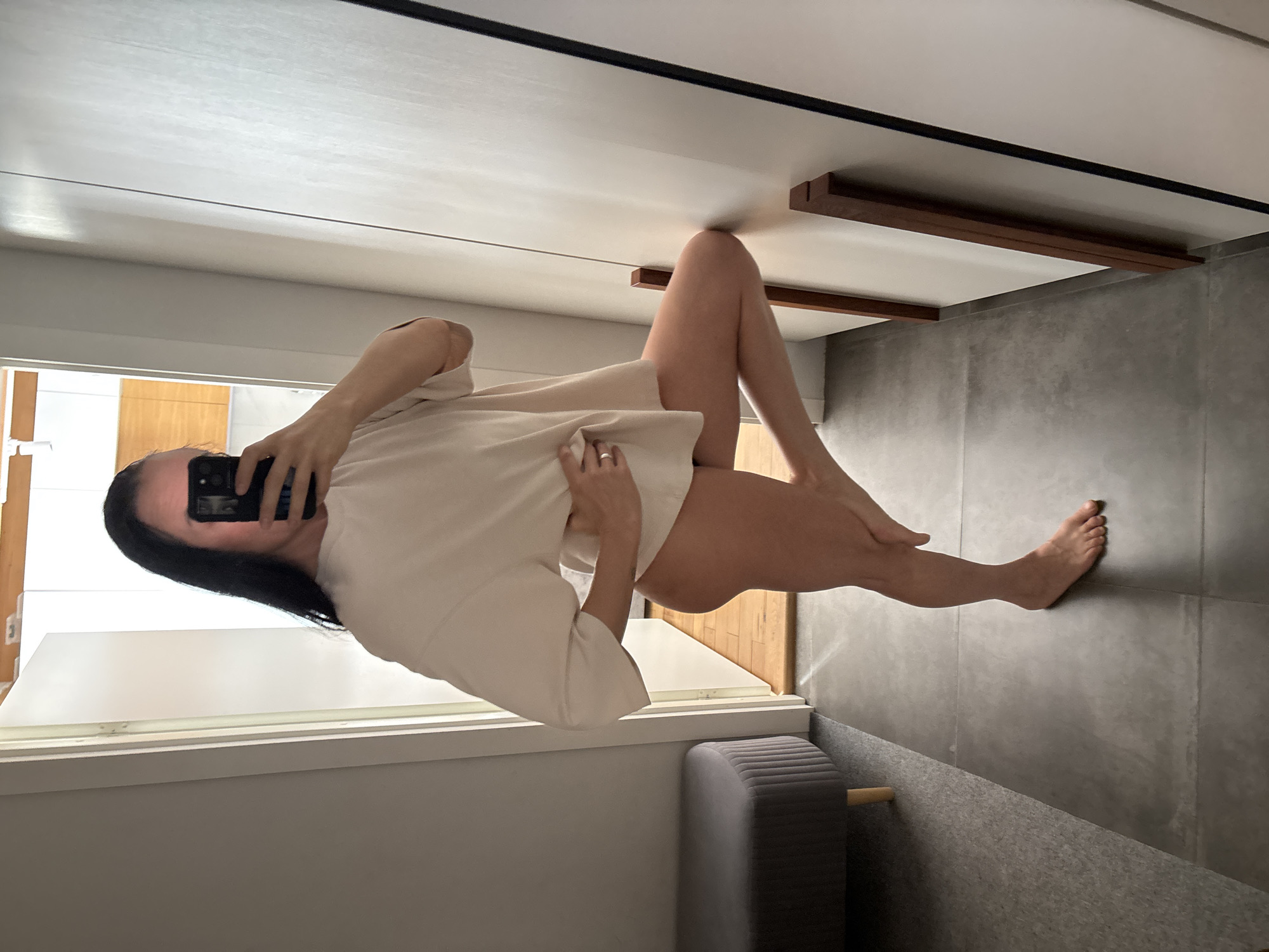 A woman with long black hair is taking a selfie in a room. She is wearing a light-colored, oversized shirt and is barefoot. She is holding her phone in her left hand. Her legs are crossed, with one foot pointing towards the right edge of the image. The background features gray tiled walls, a white window frame with natural light coming in, a light-colored wall, and a gray upholstered bench. Wood trim is visible on the ceiling. The location is in an interior setting, seemingly a room in an apartment or house, but without further context or identifying details, it is impossible to confirm the city of Chaikovsky, Russia, as the location.