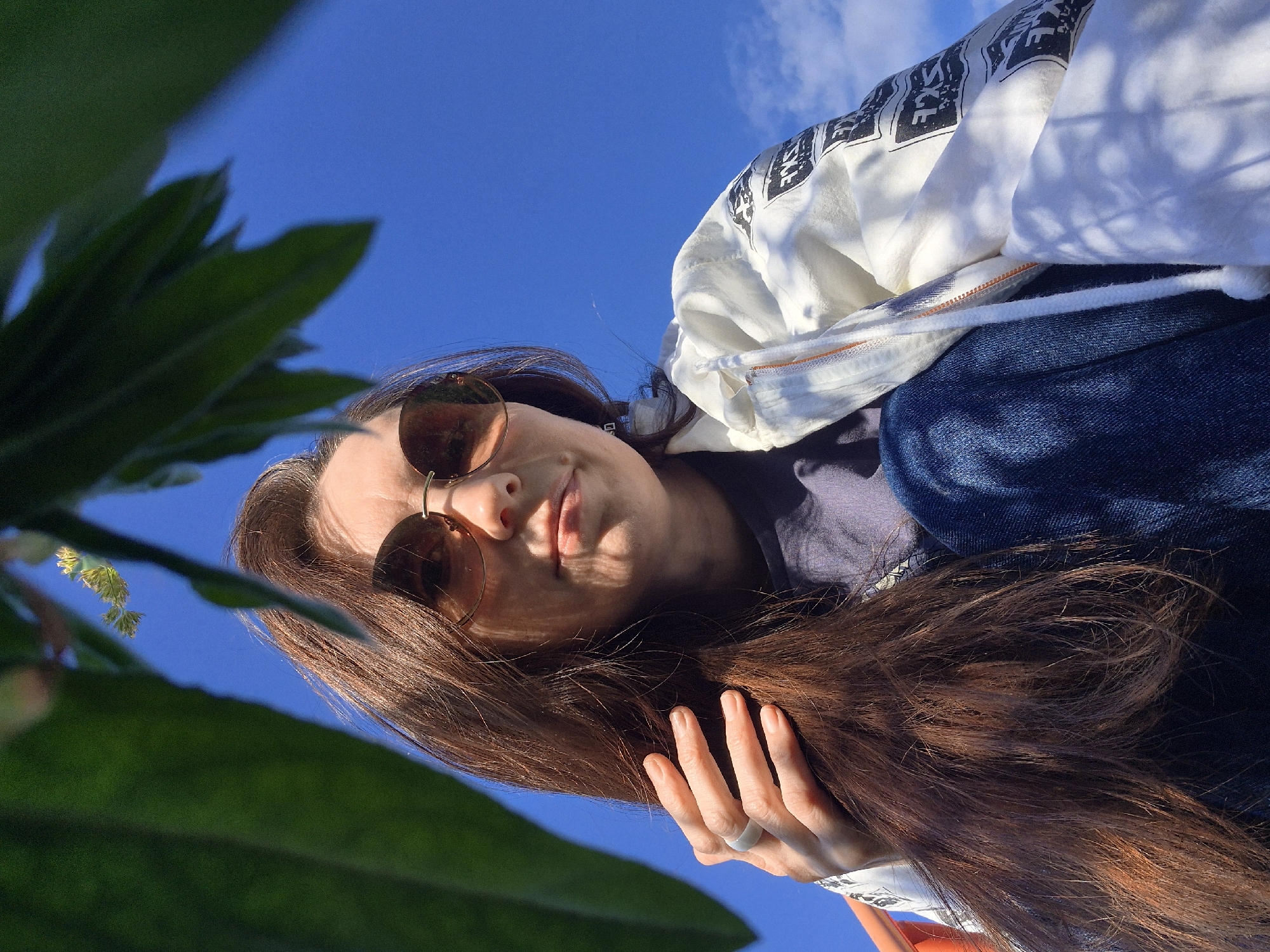 This low-angle outdoor image, rotated approximately 90 degrees counter-clockwise, captures a person looking down towards the camera against a bright blue sky. The sky, filling the upper-right portion of the frame, is clear with a few wispy white clouds. In the foreground, out of focus, are vibrant green leaves and a small cluster of light-colored buds or flowers in the lower-left.
The main subject is a young person with long, flowing dark brown hair, wearing sunglasses. The sunglasses have round, amber-tinted lenses, and their frames reflect the bright sky. The person's face is partially shadowed by the sunglasses and hair, but a calm expression with a slight, subtle smile is discernible on their lips.
They are dressed in a white hoodie or jacket featuring a repeating black graphic pattern, possibly stylized text or symbols, on the visible shoulder and sleeve area. Beneath the hoodie, a dark blue top is visible. The person is also wearing dark blue jeans. A light-colored ring is visible on one of their fingers, which is gently placed on their hair.
The bright sunlight, casting defined shadows on the person's face and clothing folds, indicates it is daytime, likely mid-morning or afternoon, with clear weather. The setting appears to be a natural outdoor environment, possibly a park or garden in Chaikovsky, Russia. The activity is a self-portrait or a photo taken from a very low perspective, emphasizing the sky and the upward gaze. No clearly legible text is visible in the image.
