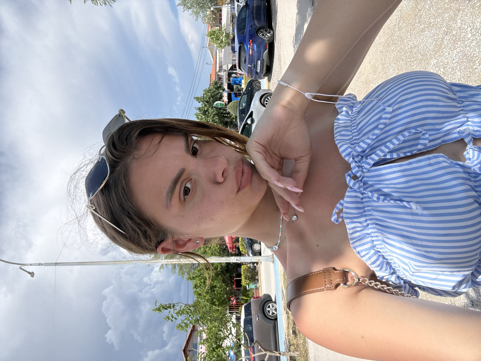 A young woman is depicted in a selfie, wearing a blue and white striped top with a brown leather and chain detail on the shoulder. She has sunglasses perched on her head, a necklace around her neck, and a bracelet on her wrist. Her left hand is near her face, and she has long, painted nails. The background shows a street scene, including parked cars, trees, buildings, and a cloudy sky. The location is in an urban environment.