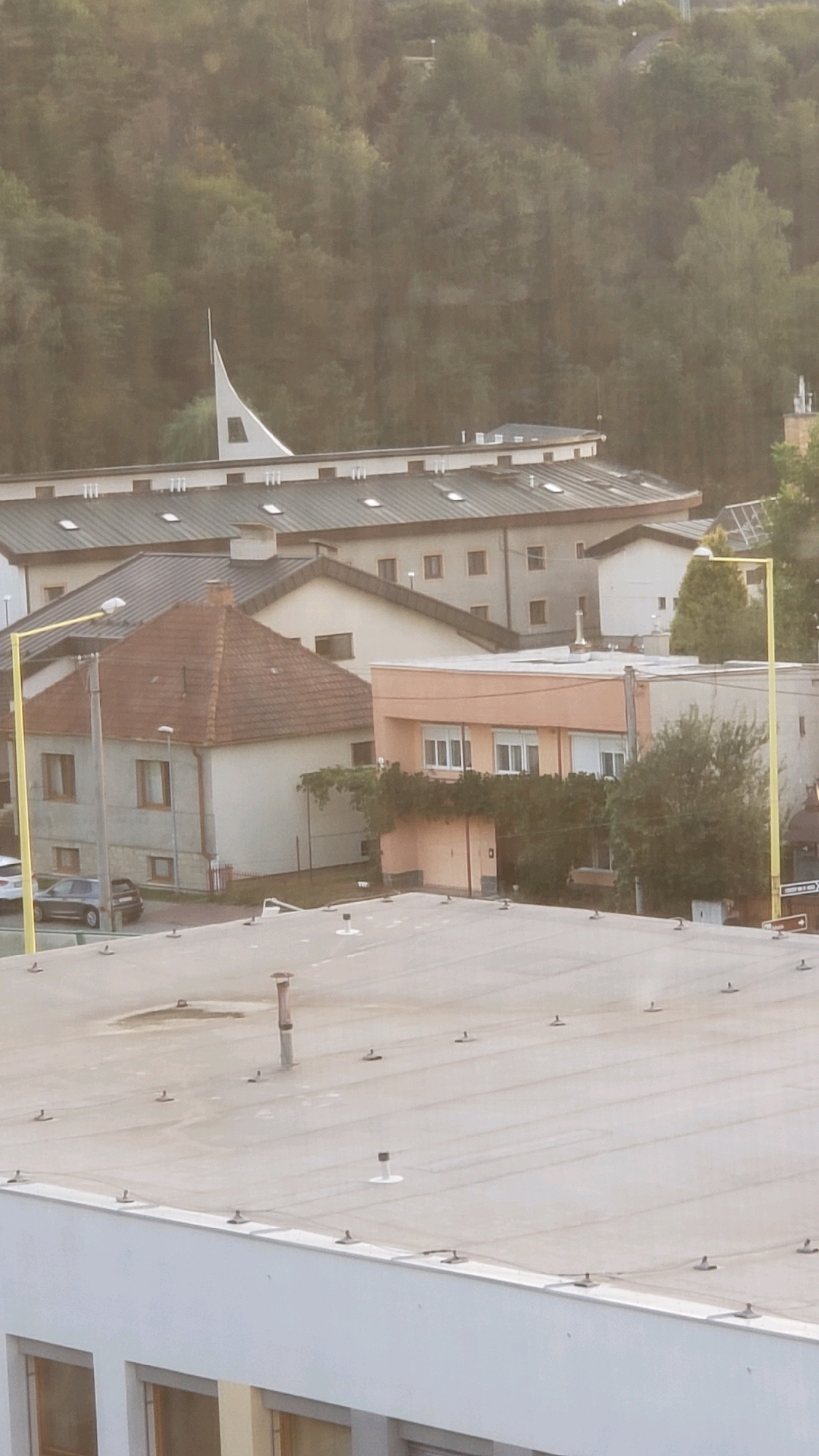 This is a high-angle, slightly hazy photograph of a suburban or semi-urban area, likely Prešov, Slovakia, taken during daylight. The foreground is dominated by a flat, light-colored rooftop with numerous small, black, T-shaped fixtures spaced evenly across its surface, suggesting it may be a building's roof, possibly a commercial or residential structure. There are also several white, cylindrical objects, some with flat tops, protruding from the rooftop.
Beyond the foreground rooftop, the scene reveals a cluster of buildings. One building, painted a light orange or peach color, has multiple windows with white frames and horizontal blinds. It is partially obscured by dense green foliage and vines. To the left of this building is a smaller, older-looking house with a red-tiled, gabled roof and light-colored walls. It features several windows with brown frames. A dark-colored car is parked on the street in front of this house, next to a yellow lamppost with a street light.
Further back, several larger buildings with dark, sloped roofs and numerous evenly spaced dormer windows are visible. These buildings appear to be multi-story structures, possibly apartment buildings or a school complex. A distinctive modern, white architectural element with a sharp, upward-pointing peak juts out from behind these buildings, set against a backdrop of a dense, green, tree-covered hillside. The overall impression is one of a developed residential or mixed-use area nestled against a natural landscape.