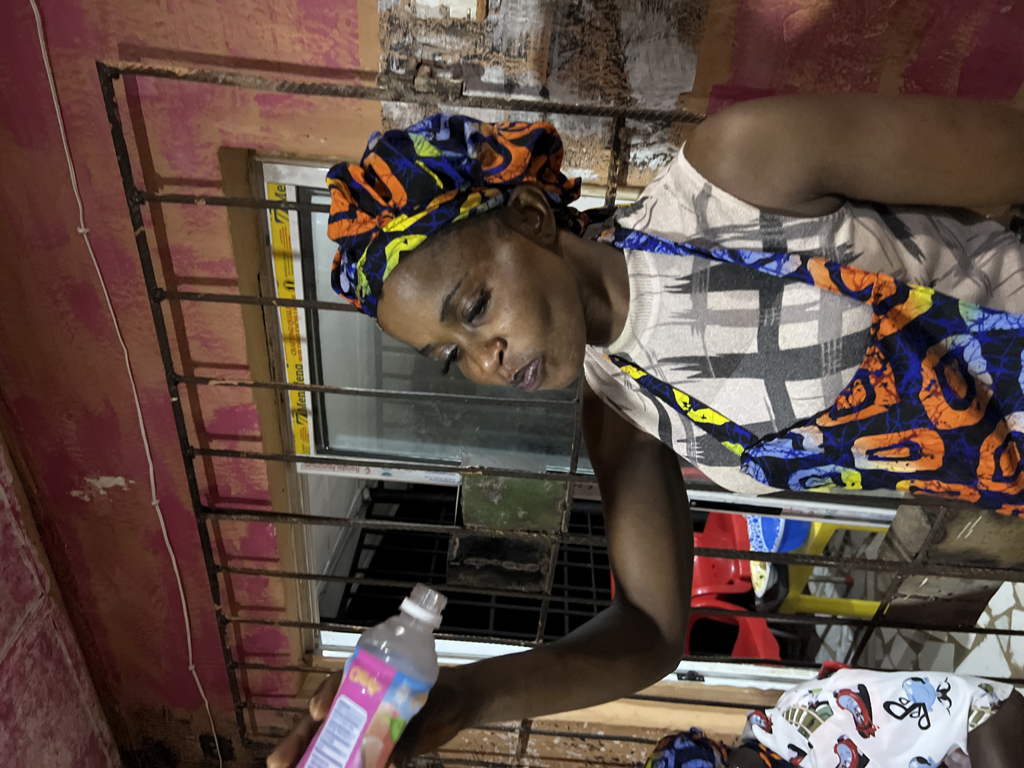 A woman, wearing a colorful headwrap and a patterned apron over a sleeveless top, is captured in a medium shot, holding a bottle of what appears to be a beverage. Her mouth is slightly open as if she is about to take a drink. The background suggests an outdoor or semi-outdoor setting, possibly a market or a street-side establishment in Nkpor, Nigeria. The wall behind her is a vibrant pink, showing signs of wear and tear. A barred window or gate runs across the middle ground, behind which a glimpse of a red plastic chair and a yellow stool can be seen, hinting at a seating area. The overall lighting suggests it is either dusk or an indoor space with artificial lighting.
