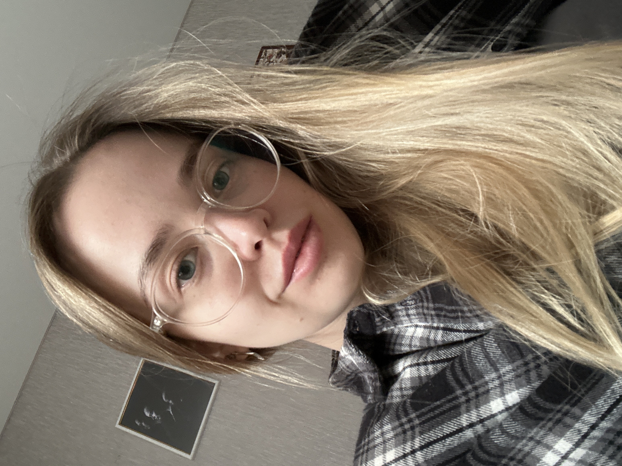 A selfie of a fair-skinned woman with long blonde hair, wearing round glasses, and a gray and white plaid shirt. The picture is taken at an angle. Behind her, a gray wall with a small framed picture is visible. The location of the picture is Chaikovsky, Russia.
