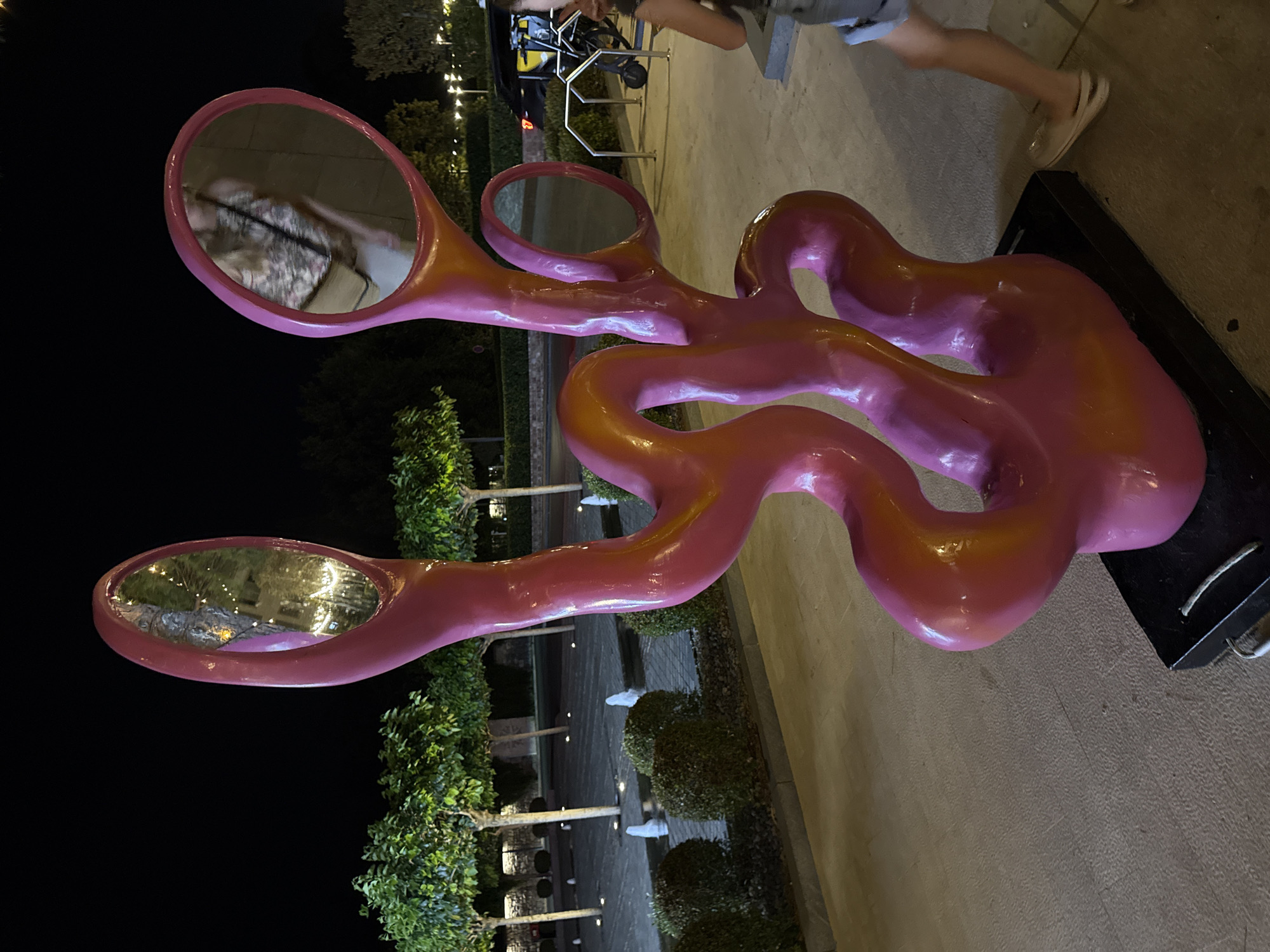 A pink and orange abstract sculpture featuring multiple oval mirrors is displayed outdoors at night, likely in a public space in Tivat, Montenegro. The sculpture's wavy, organic form is illuminated, highlighting its glossy surface. In the background, trees with uplighting and several small water fountains suggest a landscaped area, possibly a park or a promenade. A portion of a black vehicle and a bicycle rack are visible to the right, indicating an urban or developed setting. A person, mostly out of frame, is walking past the sculpture on the right side of the image, their legs and lower torso visible, wearing shorts and sandals. The sky is dark, suggesting nighttime.